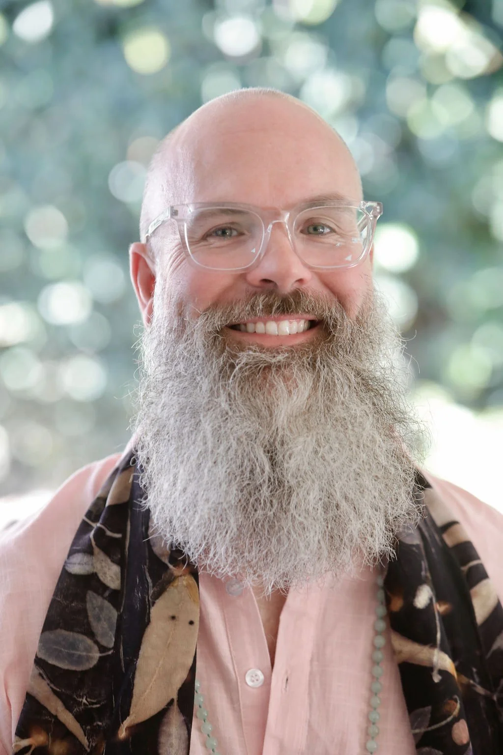 A smiling man with a large gray beard, wearing glasses, a pink shirt, and a patterned scarf, with a blurred green background.