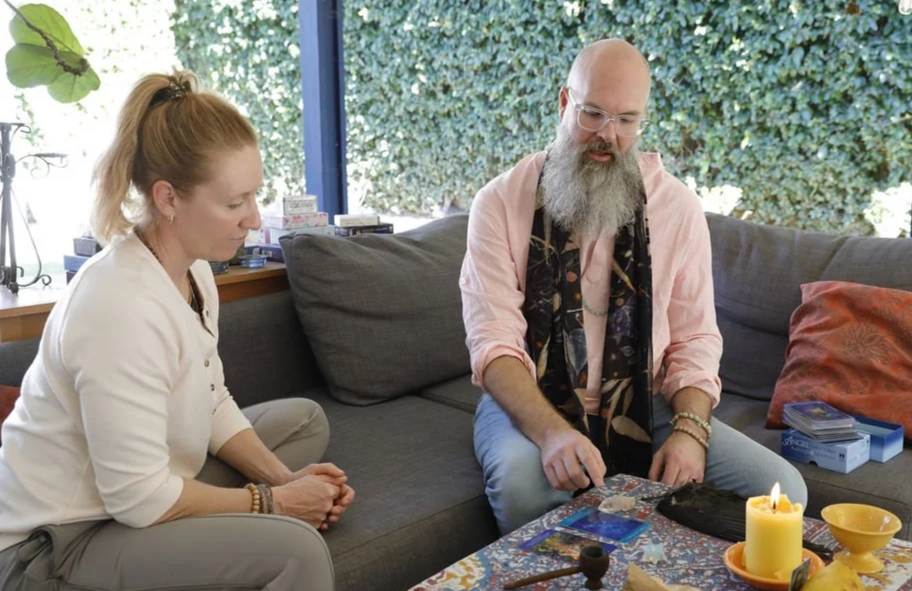 A woman and a man sitting on a couch in a sunlit room, with the woman looking thoughtfully at the man who is handling tarot cards on a colorful table.