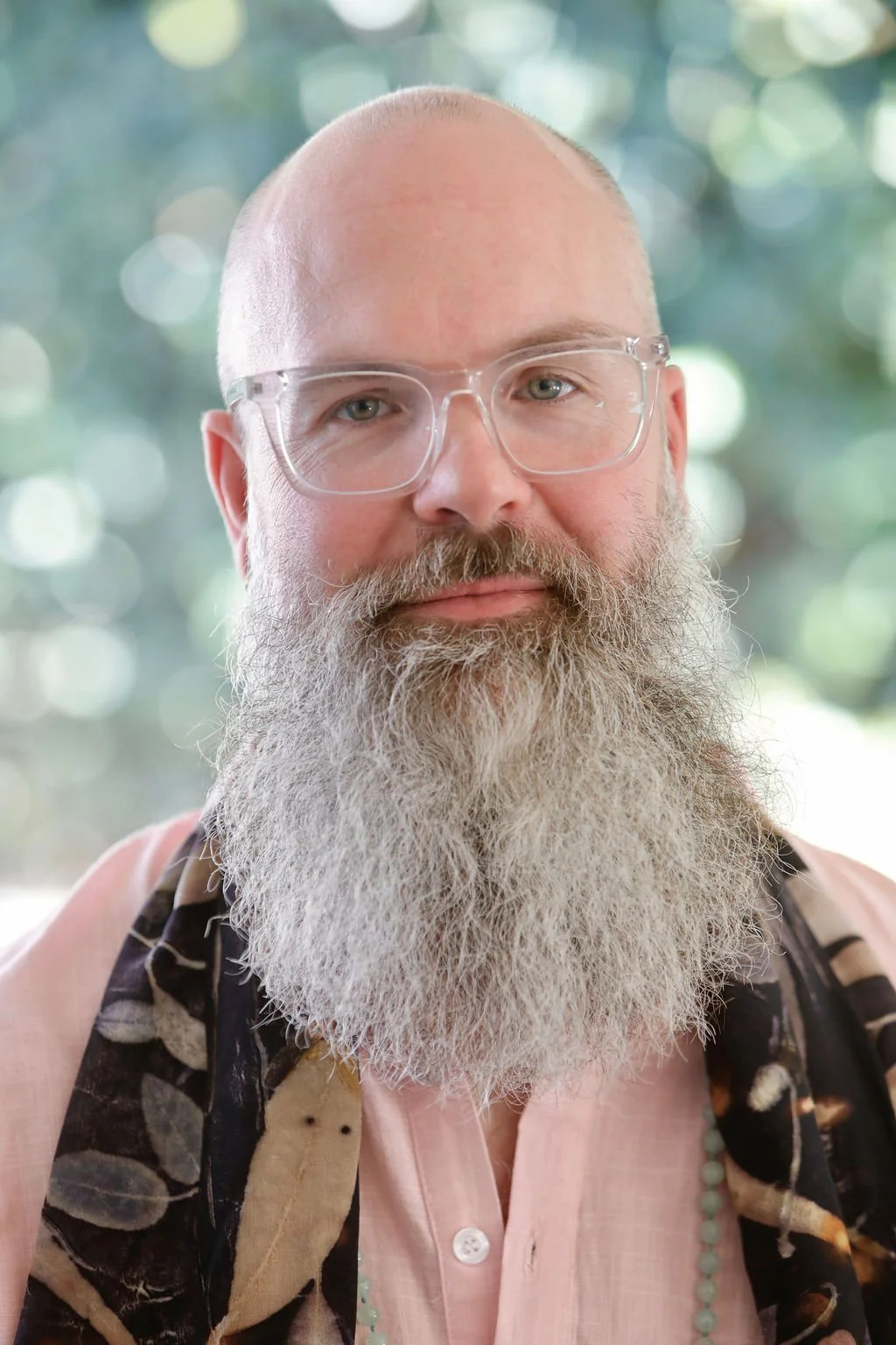 A man with a large gray beard, glasses, and a bald head looking at the camera, with a blurred green outdoor background.