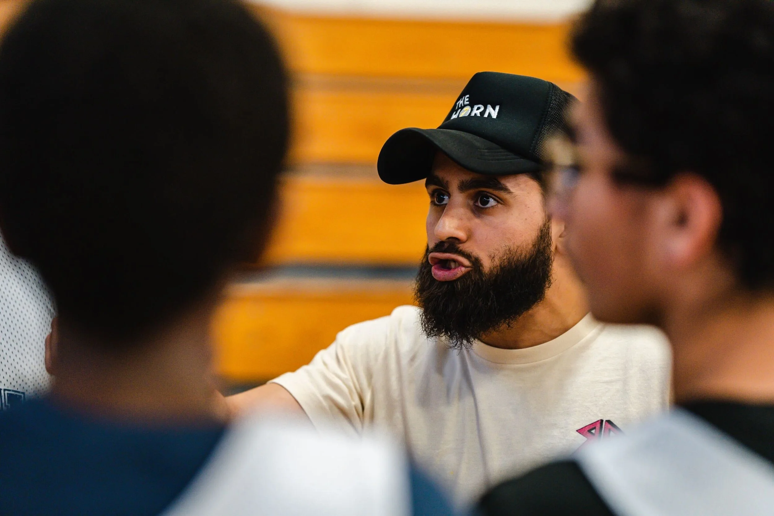 Man with a beard wearing a black cap and white t-shirt, talking passionately to a group of people in a gym-like setting.