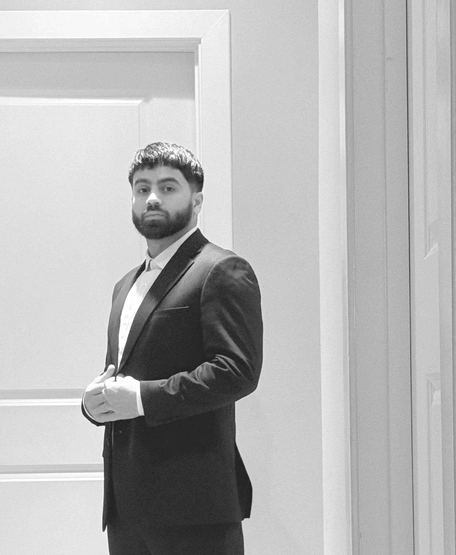 Man in a tuxedo standing in front of a plain door, looking at the camera, in black and white.