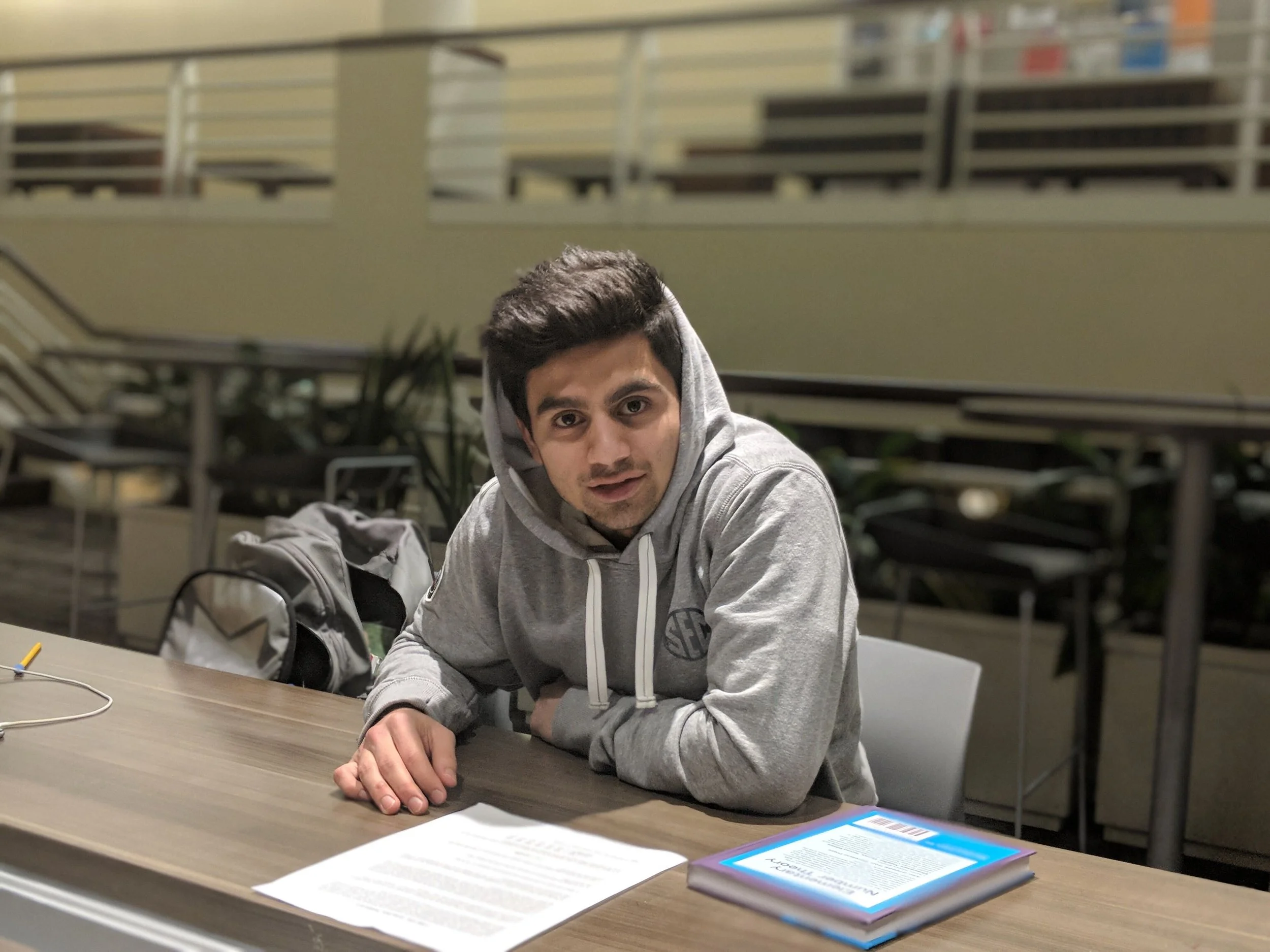 A young man with dark hair and a slight smile, wearing a gray hoodie, sits at a table with a head of papers and a tablet or electronic device in front of him. He is in an indoor setting with a railing and plants in the background.