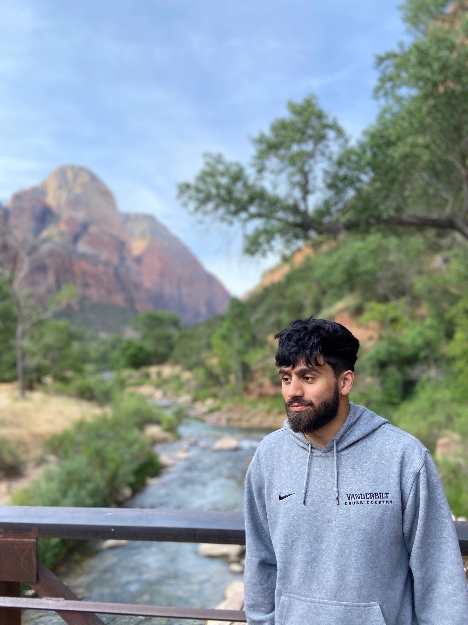 A man standing on a bridge in front of a river and lush green trees with mountains in the background