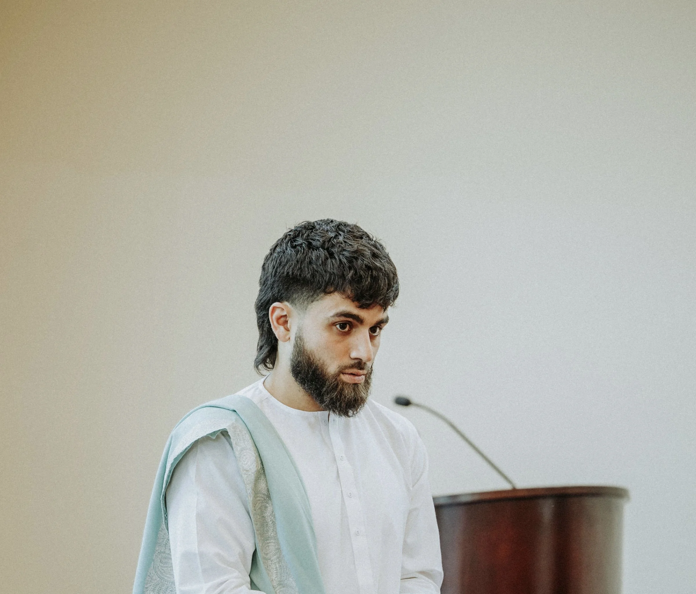 A man with dark curly hair and a beard, wearing a white shirt, stands near a wooden podium in a neutral-colored room, looking down.