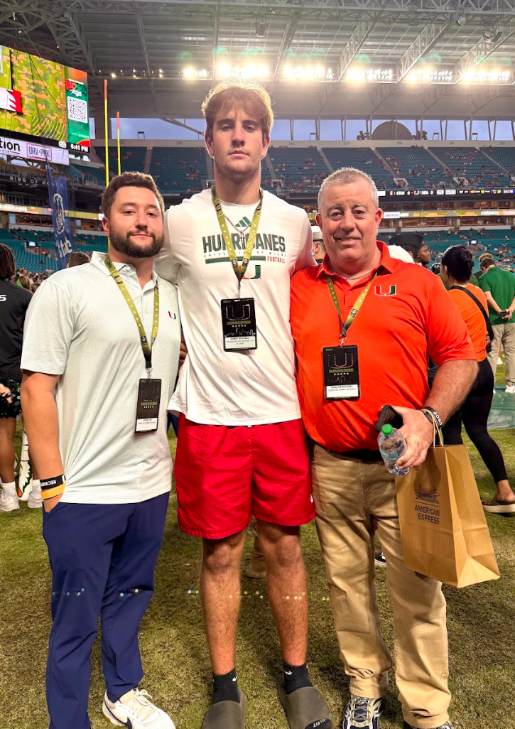 Three men standing on a football field after a game, with a stadium in the background. The man in the middle is wearing a white 'Hurricanes' football t-shirt and red shorts, with a lanyard and a badge around his neck, and is taller than the others. The man on the right is wearing an orange polo shirt with the University of Miami logo, khaki pants, and is holding a water bottle and a brown paper bag. The man on the left is dressed in a white polo shirt, navy pants, and also has a lanyard with a badge. They are smiling and posing for the photo.