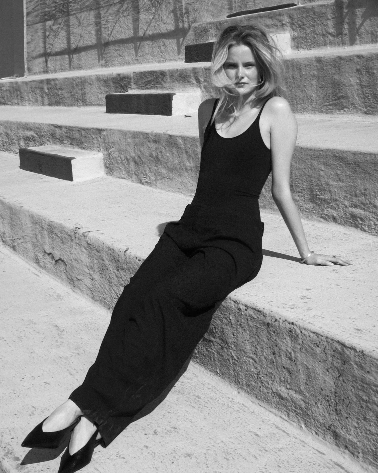 Black-and-white fashion portrait of a female model with blonde hair, sitting on concrete steps wearing a black tank top and wide-leg pants, shot in bright sunlight with geometric shadows in the background.