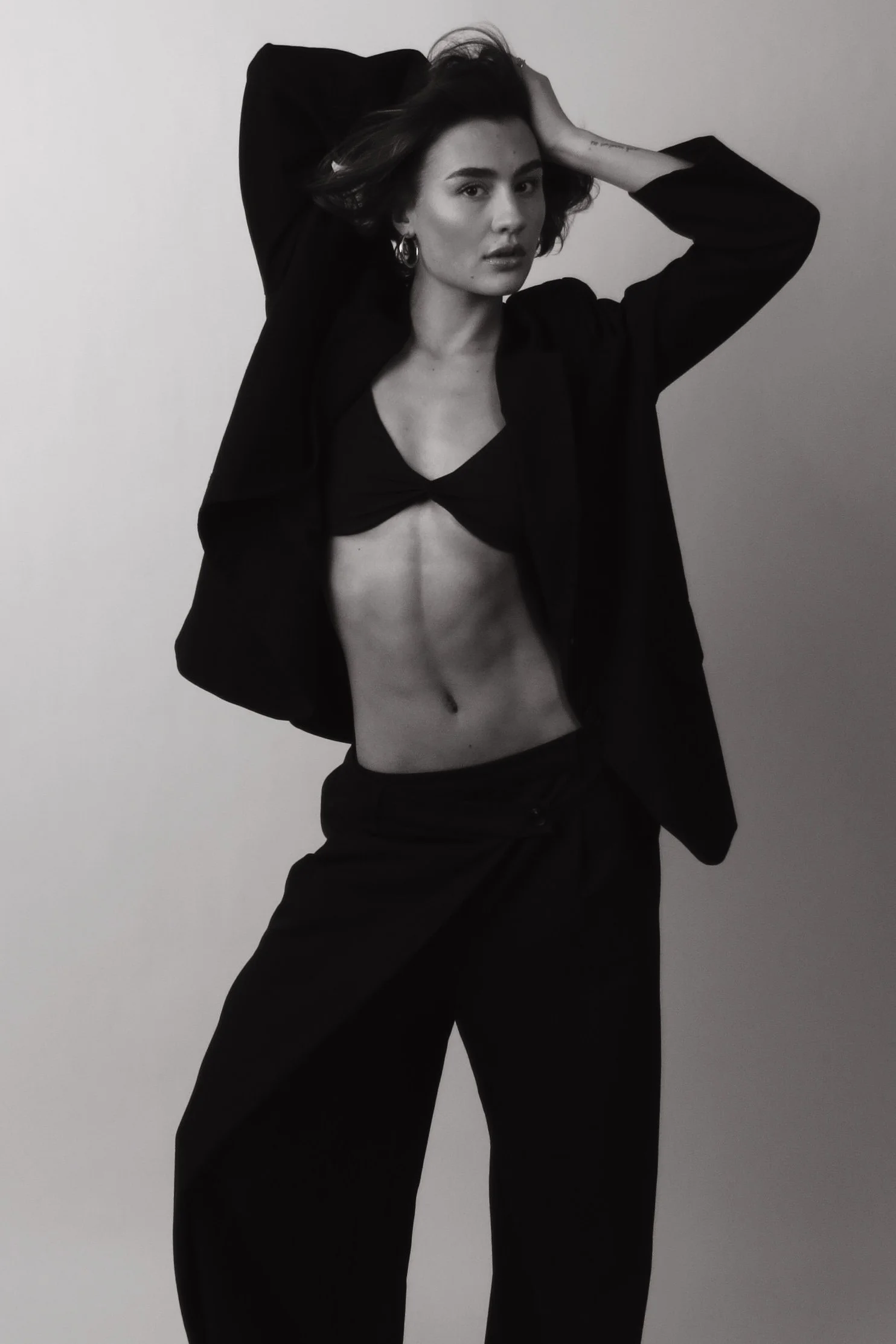 Zwart-wit foto van een vrouw met kort haar, die een zwarte blazer en broek draagt, haar linkerarm omhoog en haar rechterarm over haar hoofd, poseert tegen een effen muur.