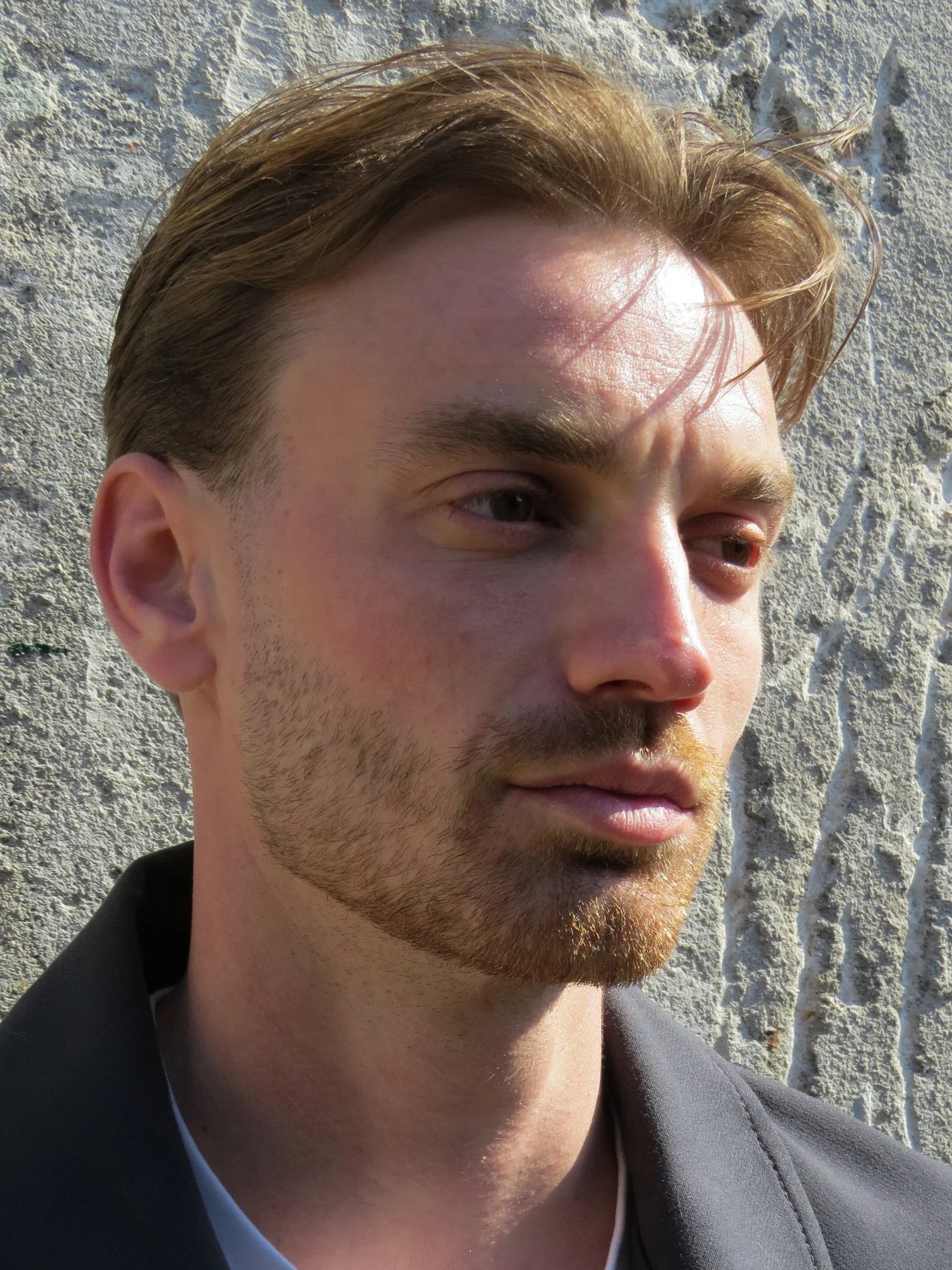 Close-up van een man met lichtbruin haar en een baard, gekleed in een donker pak met een witte shirt, zittend voor een witte bakstenen muur.