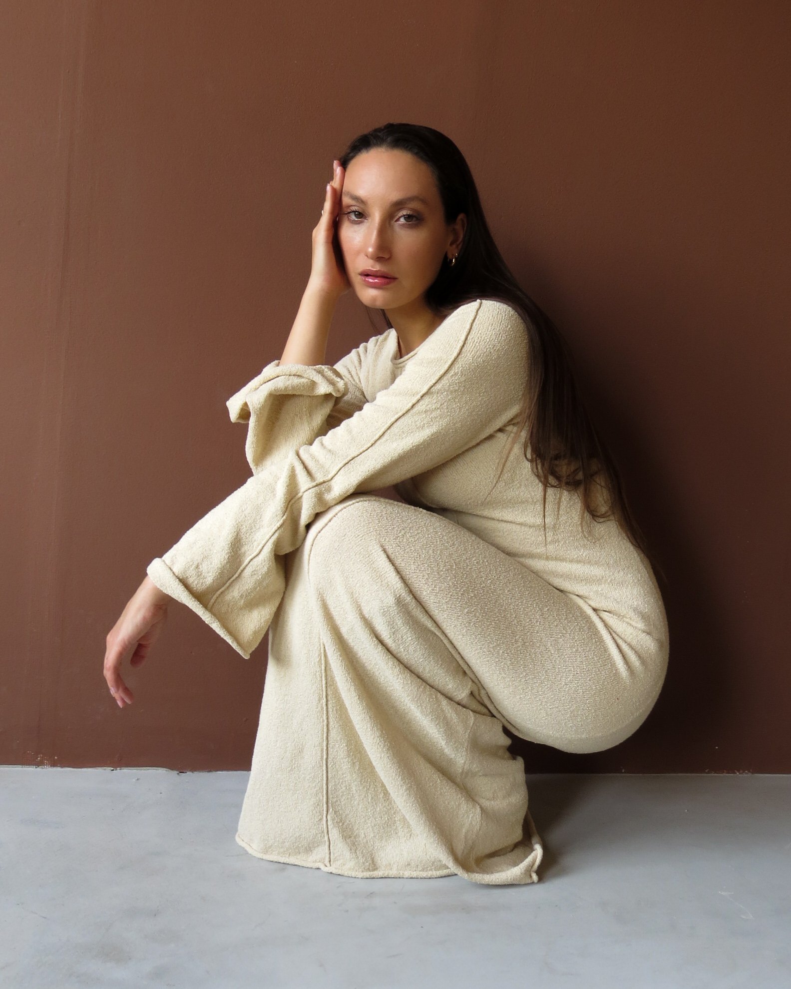 Een vrouw in een beige jurk zit op de grond voor een bruine muur, haar hoofd rust op haar hand en haar blik is richting de camera.