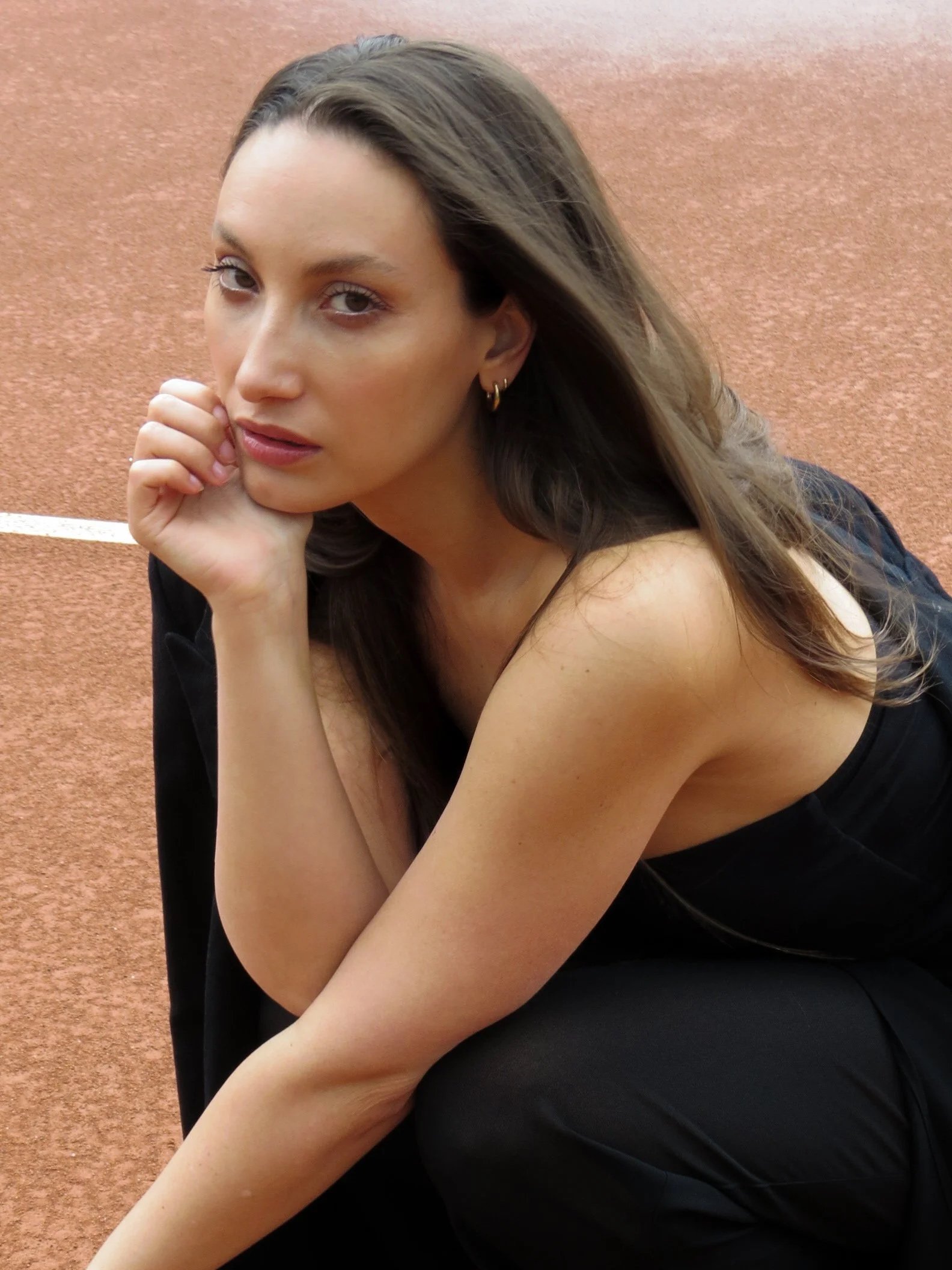 Close-up van een jonge vrouw met lang bruin haar, die op een sportterrein met een zandachtige ondergrond zit, en rust met haar hoofd op haar hand.