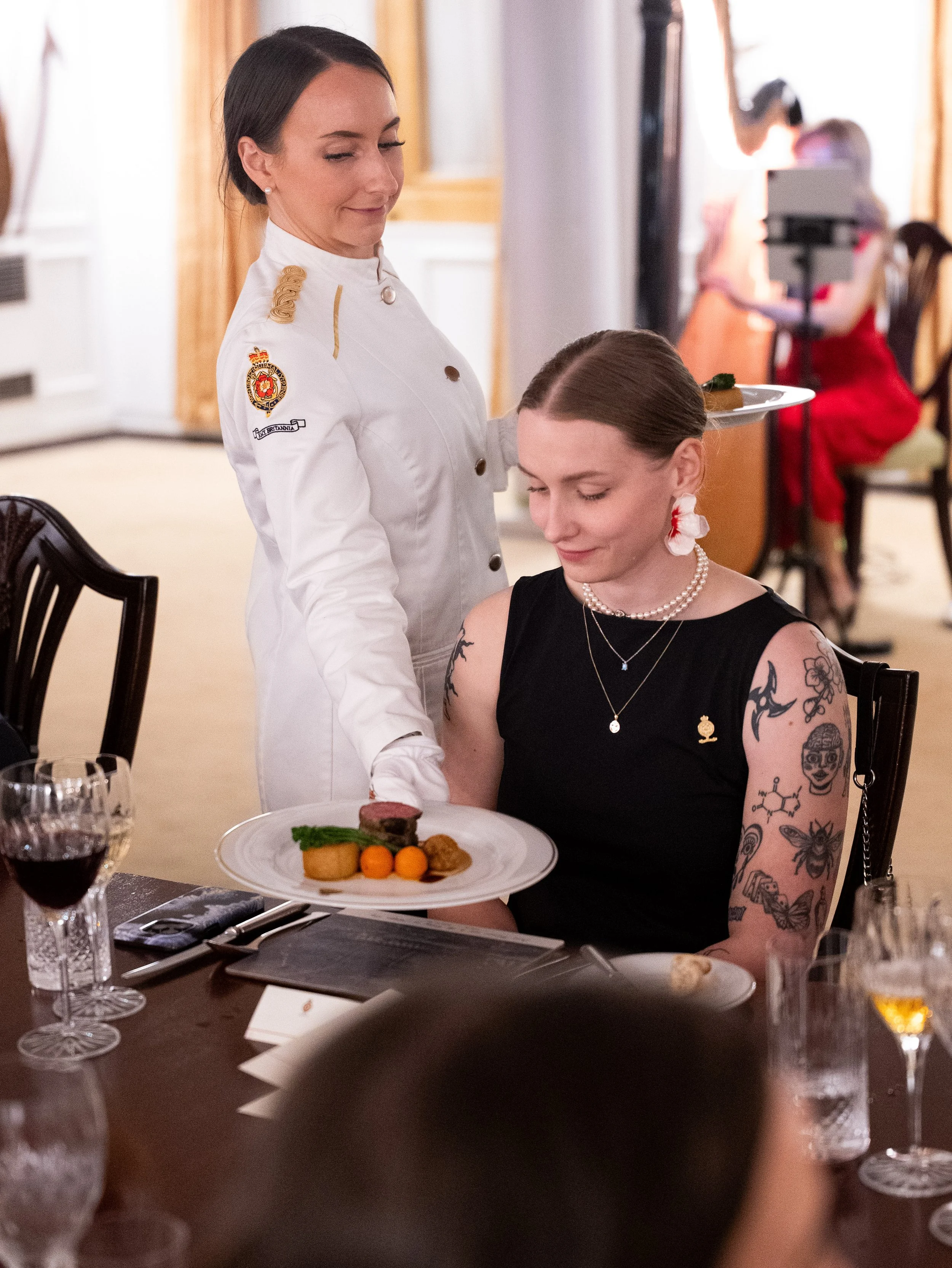 A female chef serving a plated dish to a seated woman with tattoos in a formal dining setting.
