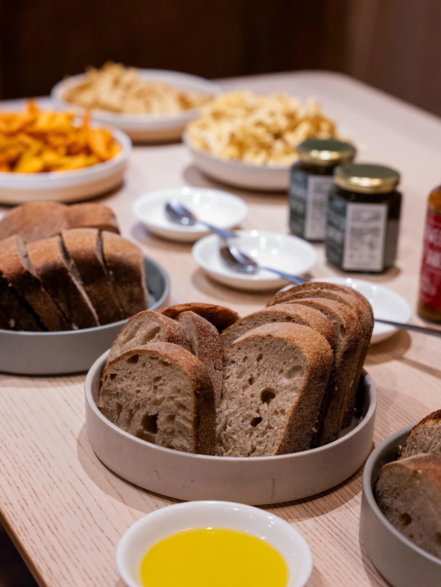 Sliced bread in a white bowl on a wooden table with bowls of pasta, jars, and small plates in the background.
