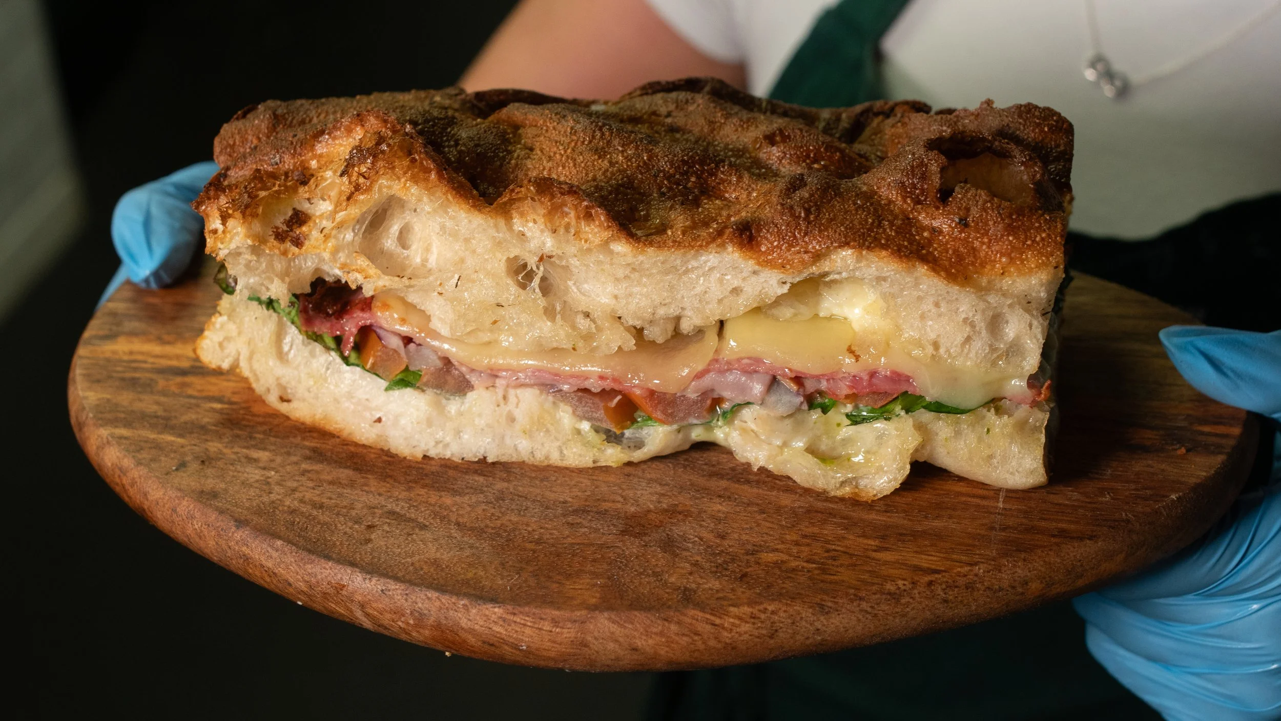 A large Italian sandwich with toasted bread filled with cheese, ham, and greens, being held on a wooden serving board by someone wearing blue gloves.