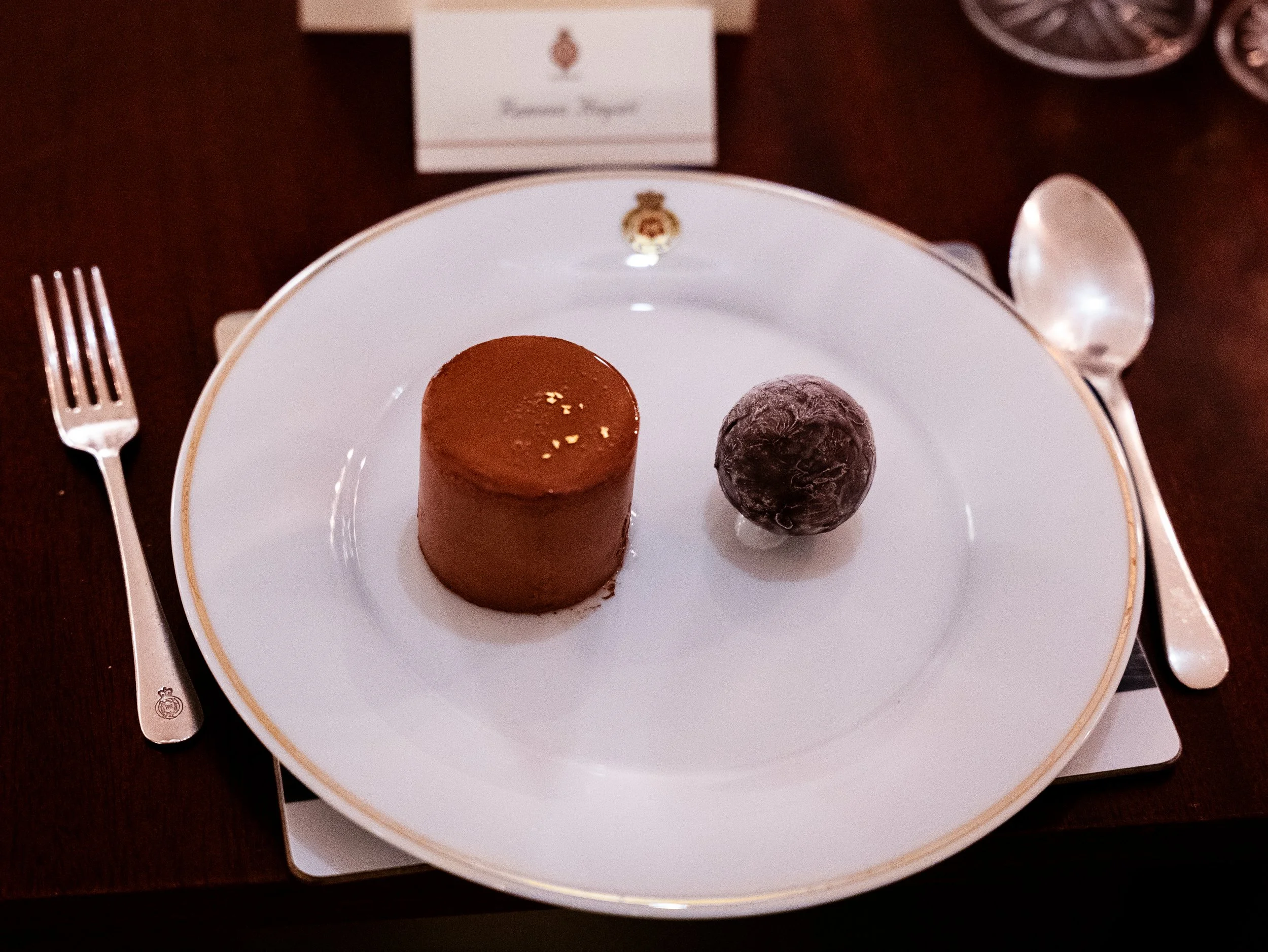 A plate with a chocolate mousse and a chocolate-covered truffle. The plate is on a table set for a meal with a fork on the left and a spoon on the right.