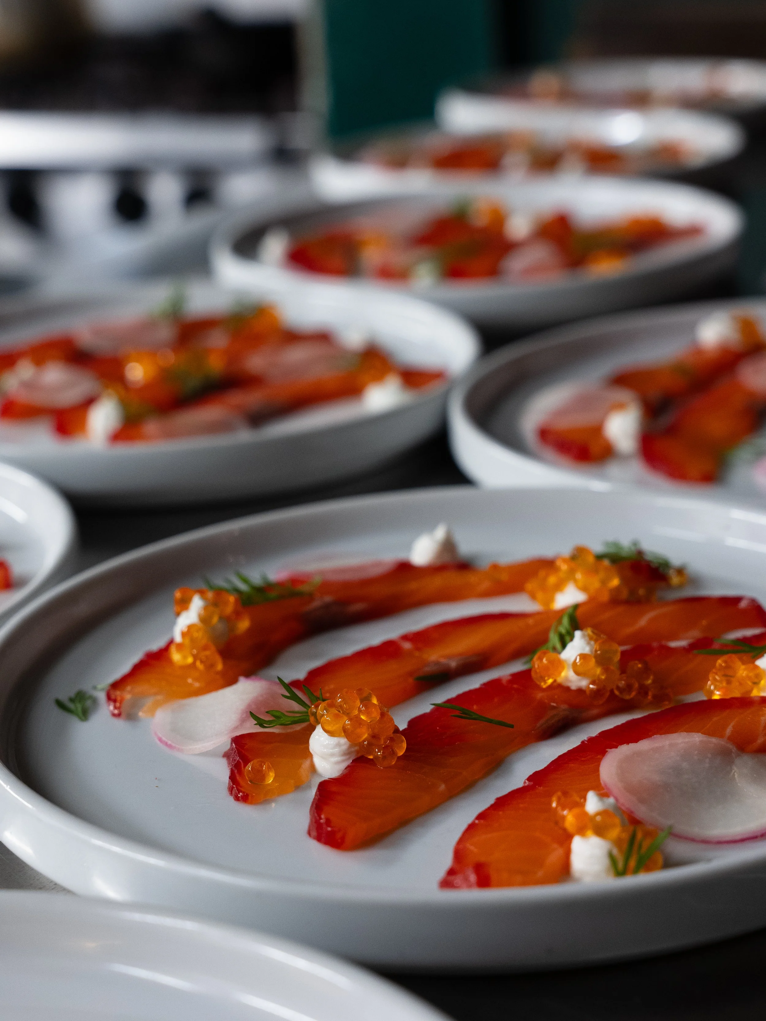 Several plates with slices of smoked salmon, radish slices, dollops of cream, and garnishes, arranged on a dark dining table.