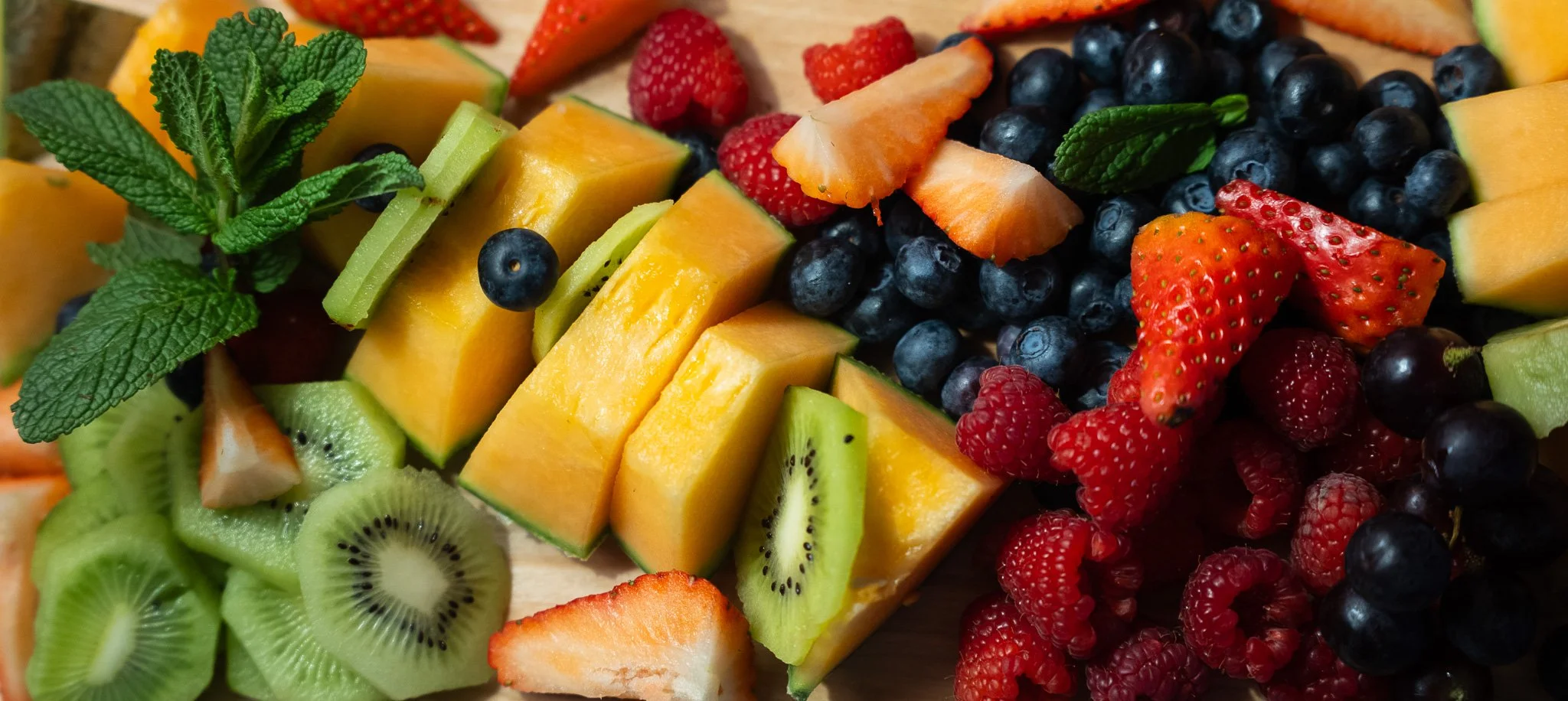 Close-up of an assortment of fresh sliced fruits, including strawberries, blueberries, raspberries, kiwi, cantaloupe, honeydew, and slices of mango, garnished with a sprig of mint.