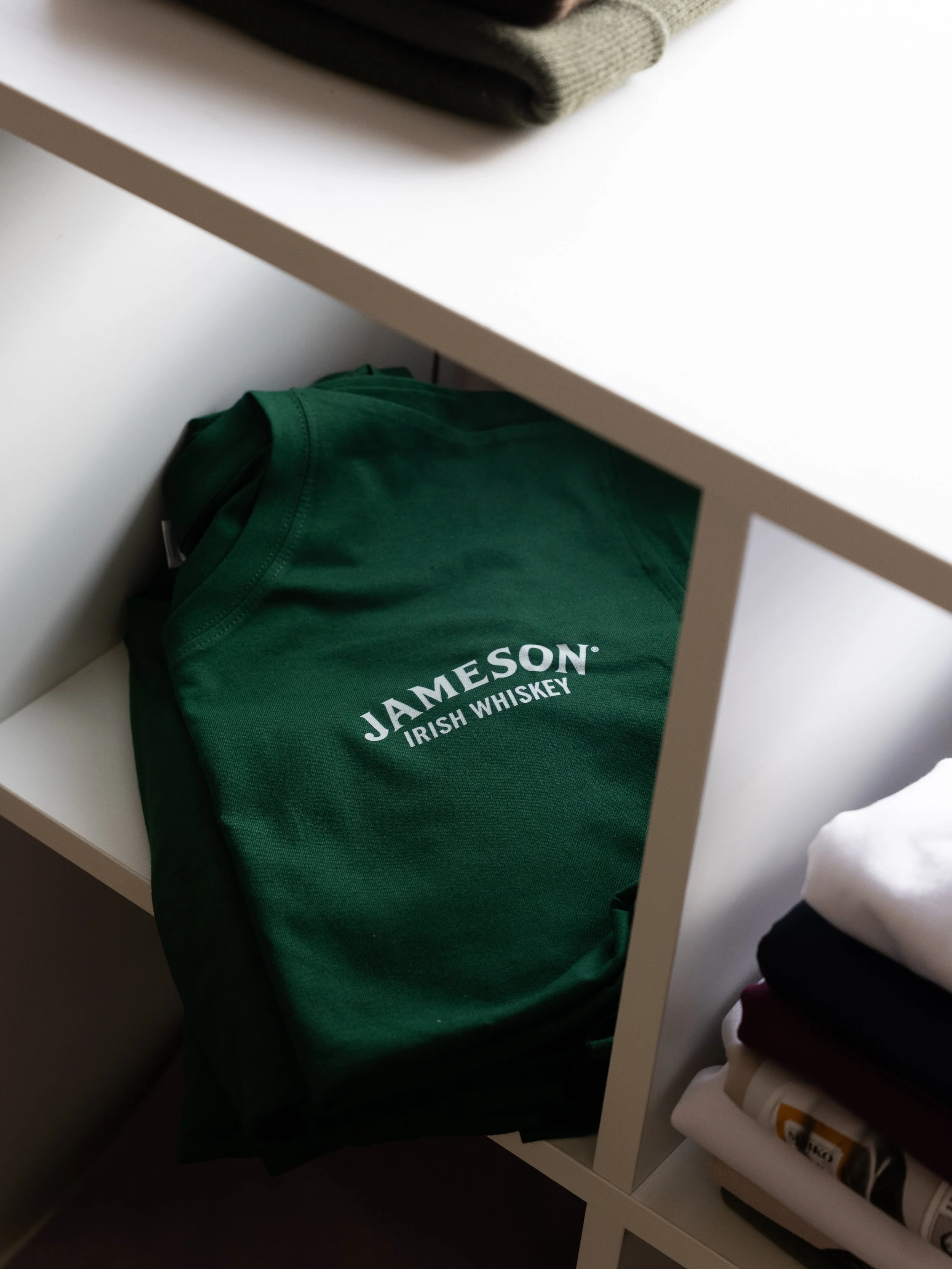 A green t-shirt with 'Jameson Irish Whiskey' logo folded and placed inside a white shelf.