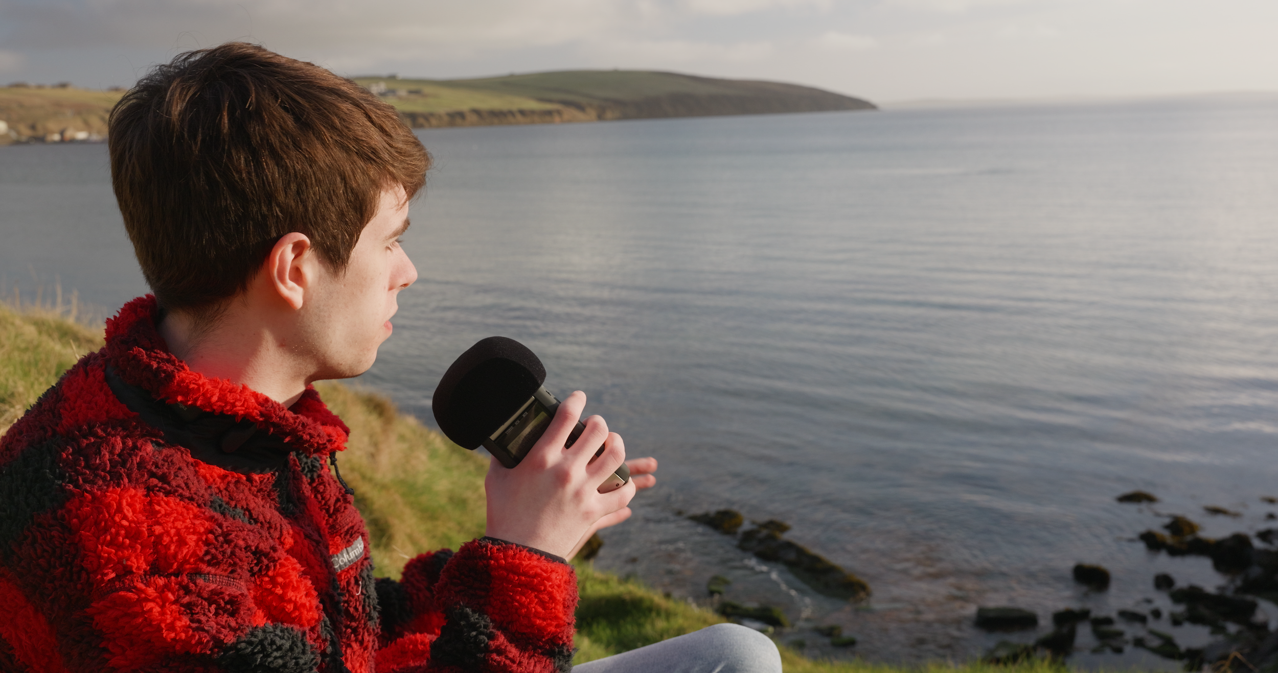 Young man in a red and black fleece jacket sitting on a grassy hill near the coast, holding a black microphone, with a calm body of water and distant land in the background.