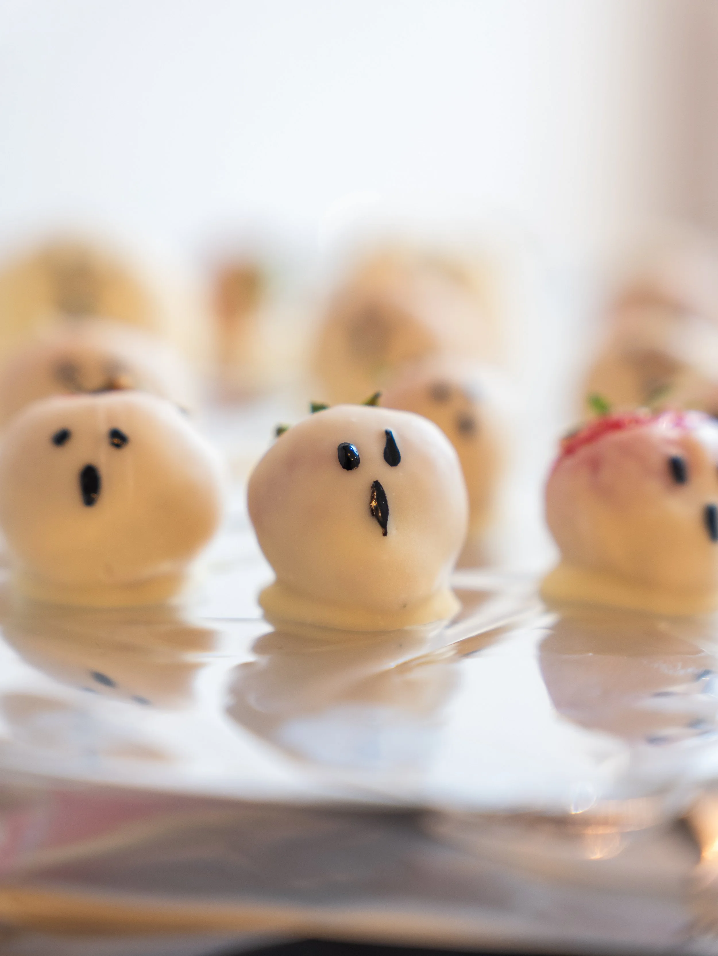 White chocolate strawberries with black decorative eyes and mouths, some with small green leaves on top, arranged on a reflective silver surface.