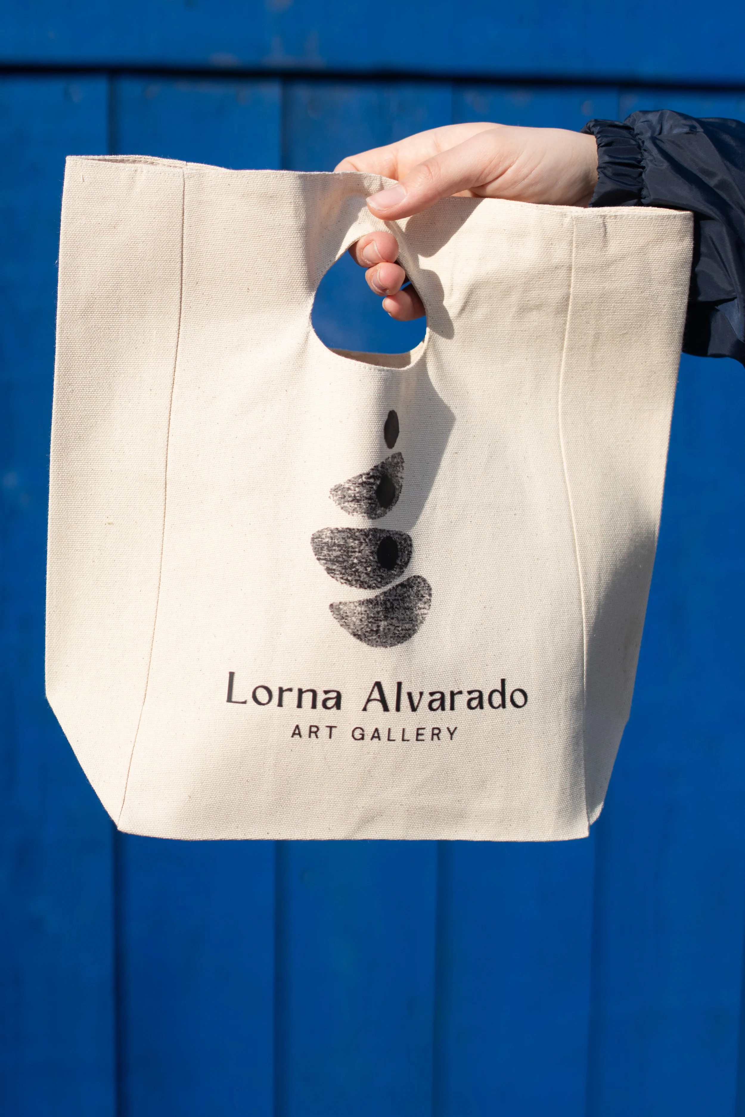 Person holding a beige tote bag with a minimalist black footprint design and the text 'Lorna Alvarado ART GALLERY' printed on it, standing in front of a blue wall.