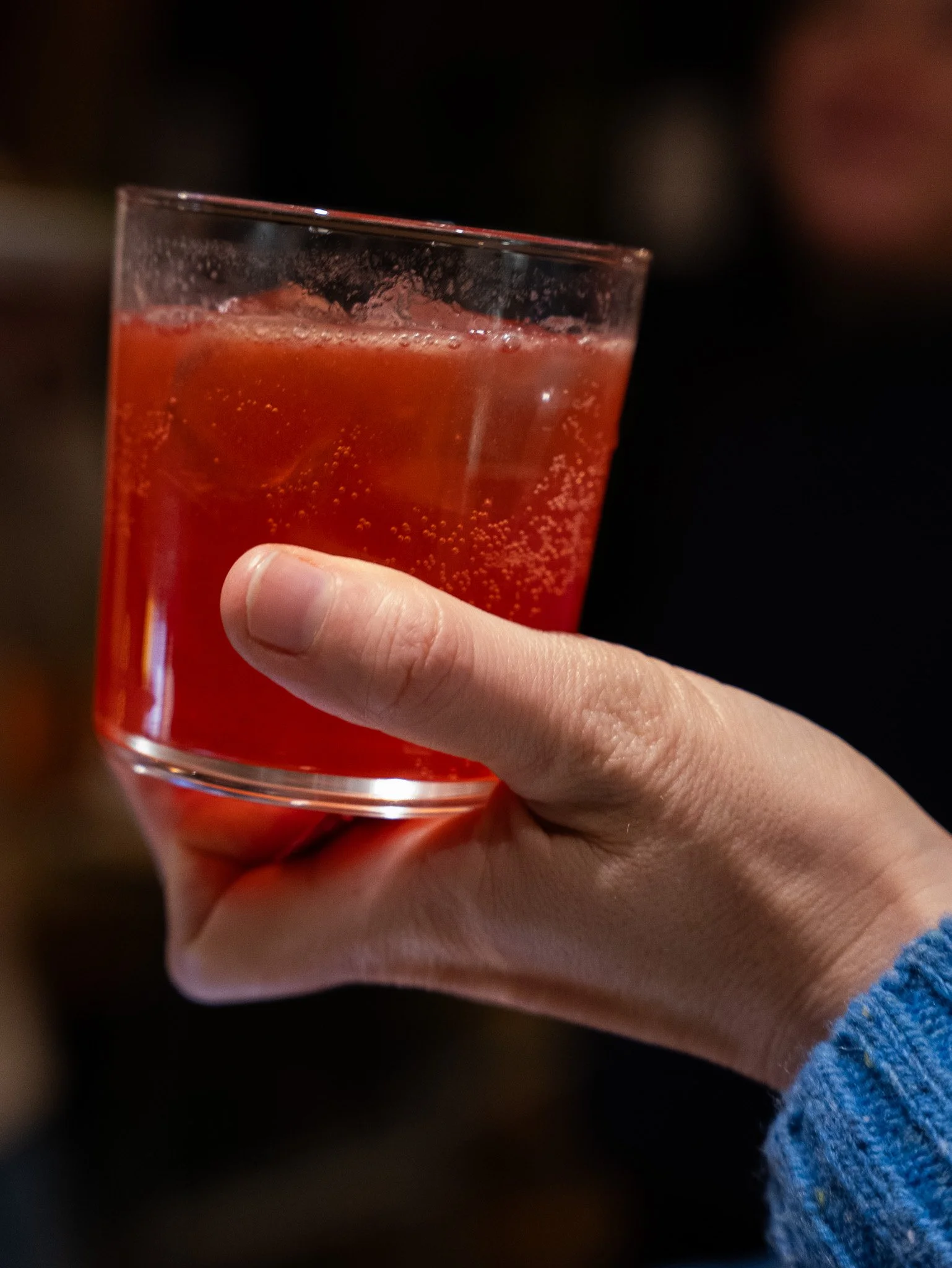 A hand holding a glass of red fizzy drink with ice cubes, against a dark background.