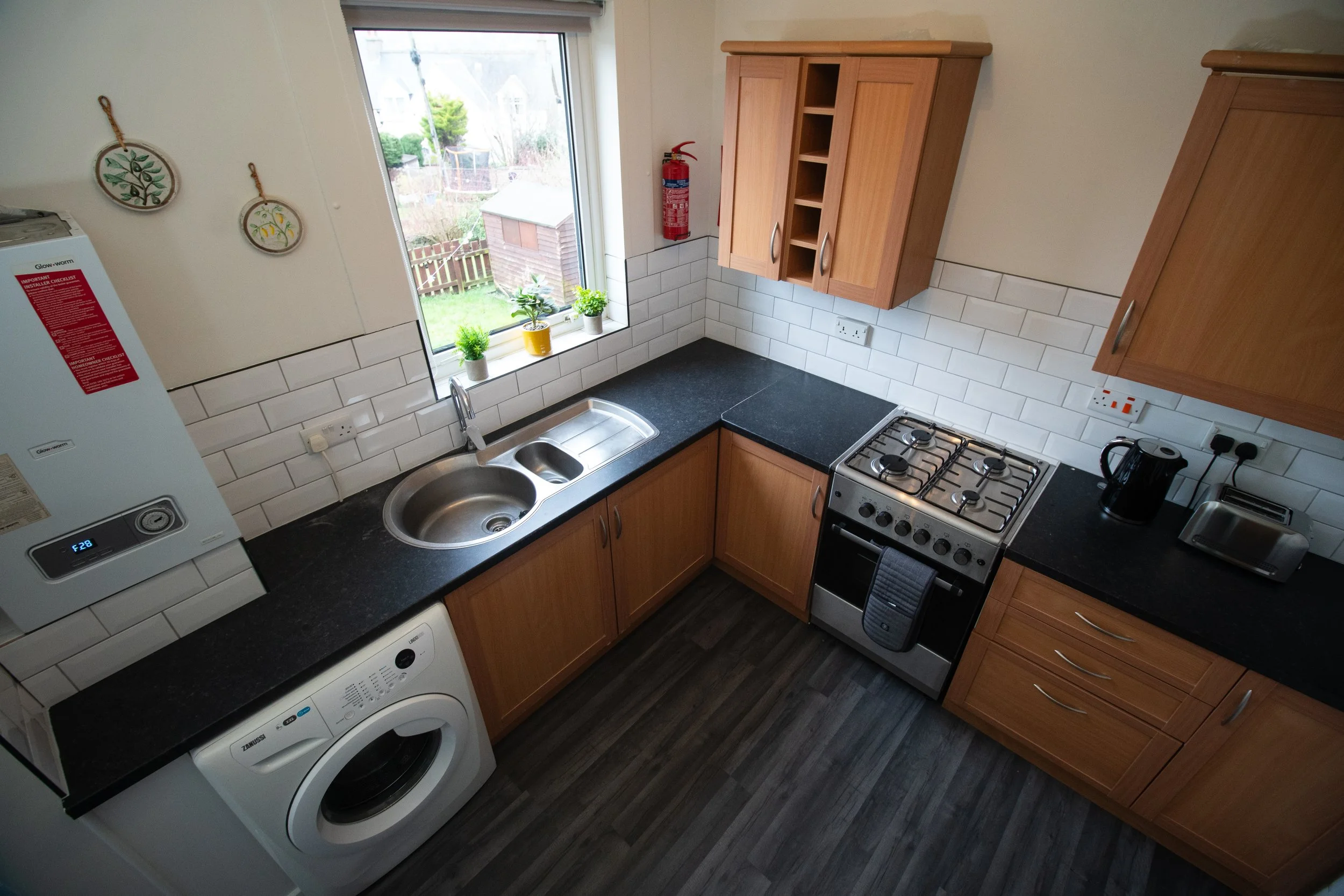 A kitchen with wooden cabinets, black countertops, a stainless steel oven, a washing machine beneath the counter, a black kettle, toaster, and electrical outlets. There is a window above the sink with small plants on the windowsill and a view of a garden outside.