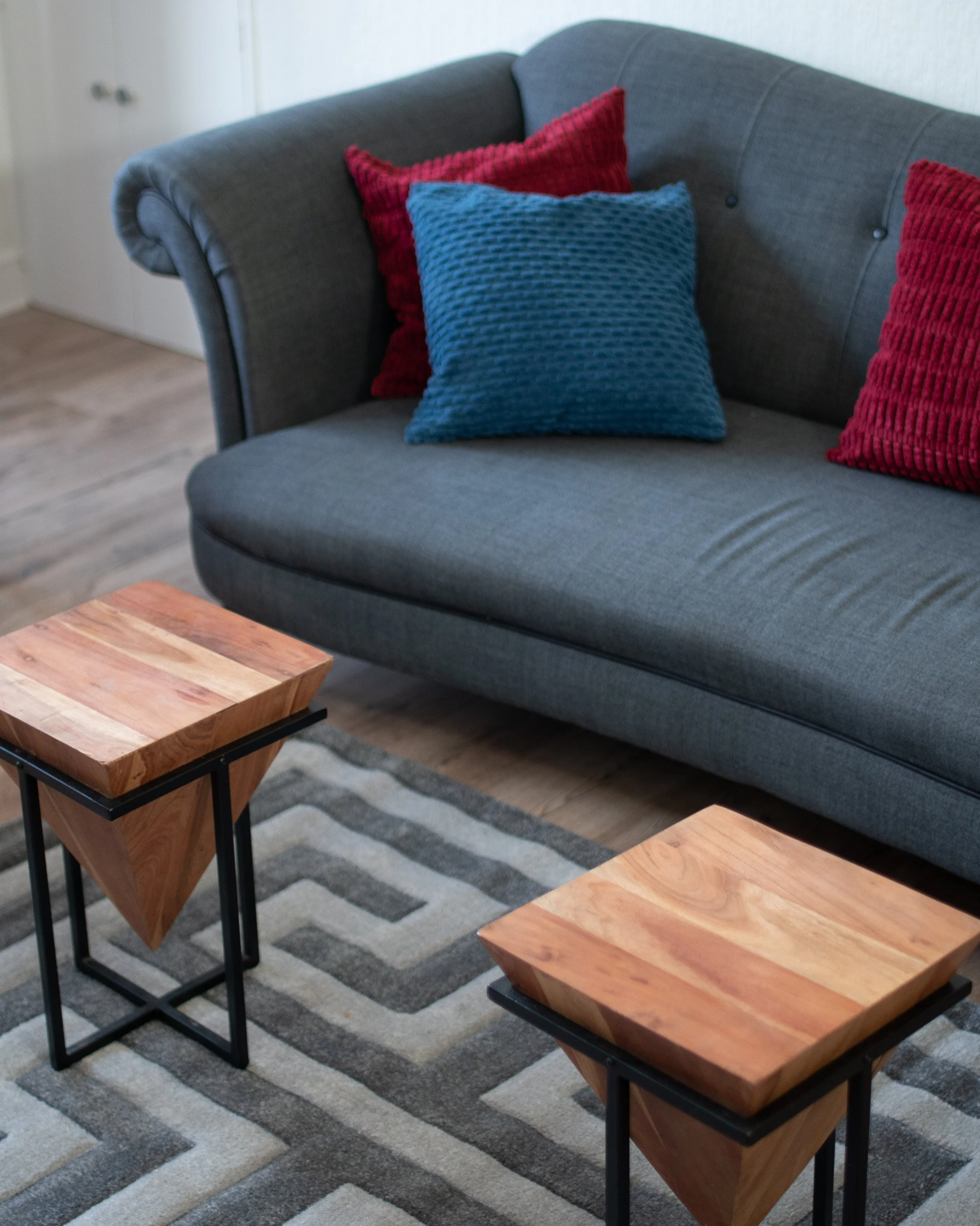 A living room with a gray sofa, red and blue textured pillows, a patterned area rug, and two wooden side tables with black metal frames.