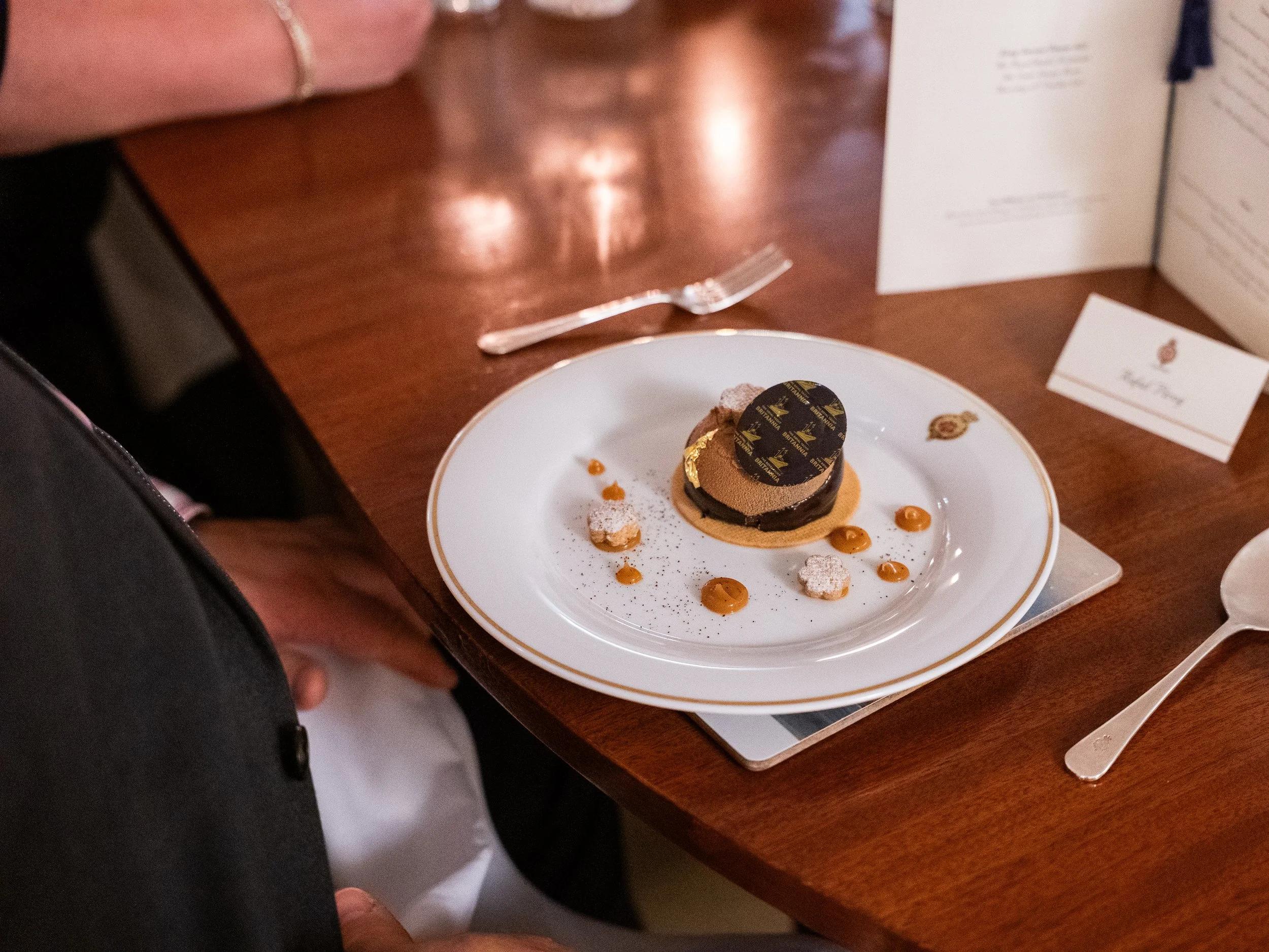 A dessert presented on a white plate with gold trim, featuring a chocolate mousse topped with a round chocolate disk with gold accents. The plate is decorated with small dollops of caramel sauce, powdered sugar, and small white meringue cookies dusted with powdered sugar. The dessert is set on a wooden table near a fork, a spoon, a menu, and a place card.