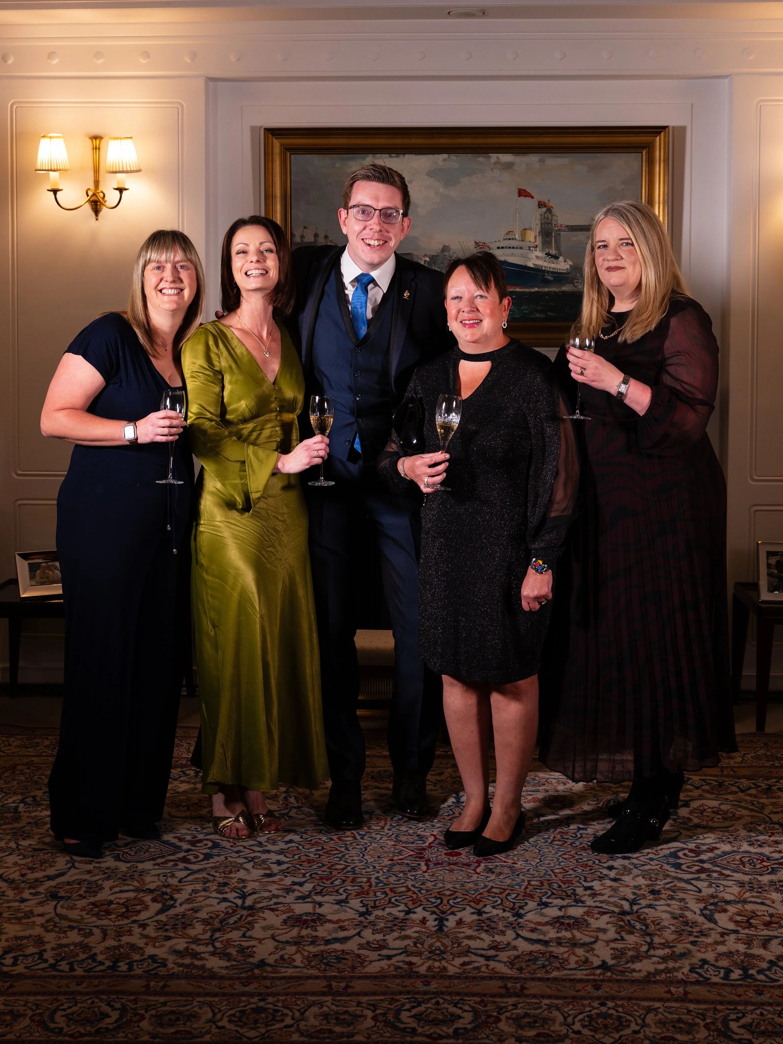Six adults dressed in formal attire, holding wine glasses, standing together in a well-decorated room with a painting of a ship and a cozy lamp on the wall behind them.