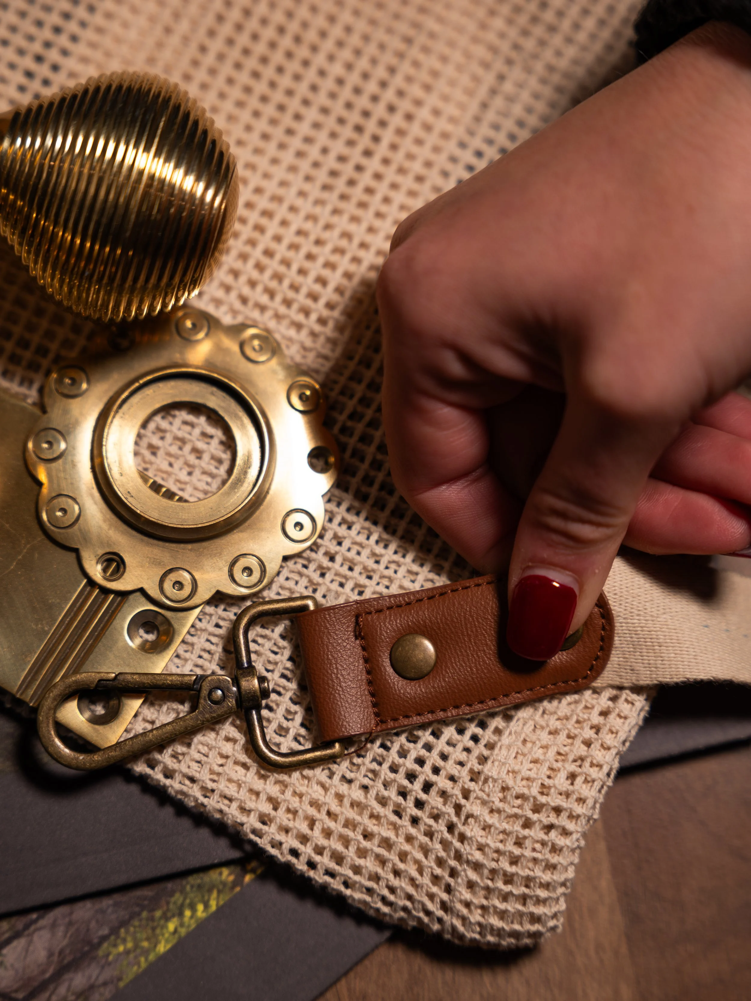 Close-up of a person's hand with red nail polish, holding a leather strap with a metal snap button. Nearby are various metallic camera parts including a lens and gear, all placed on a woven fabric surface.