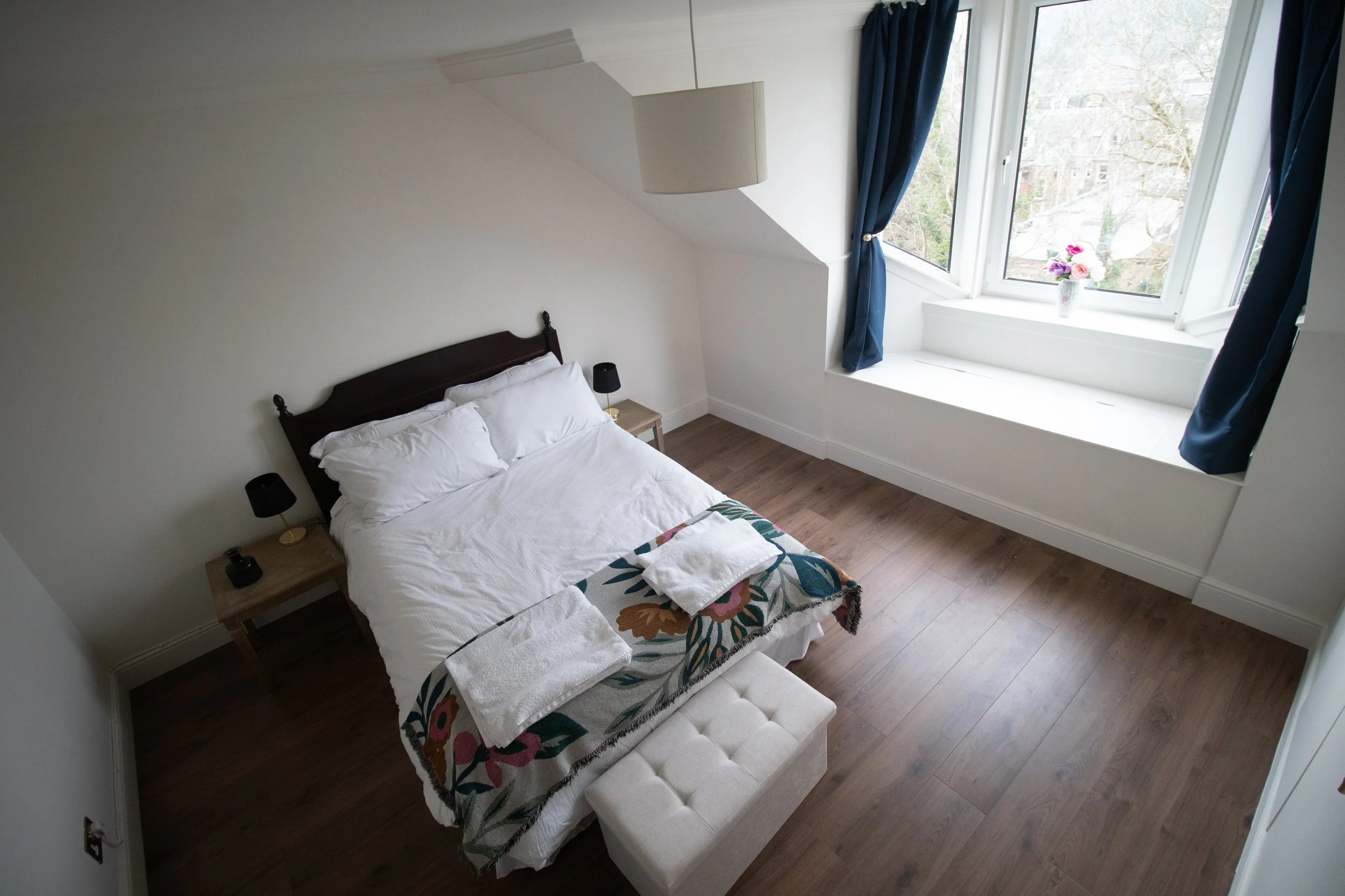 A bedroom with a double bed covered in white bedding, two nightstands with black lamps, a floral blanket at foot of bed, a window with blue curtains and a flower vase, and a cream-colored ottoman in front of the bed.