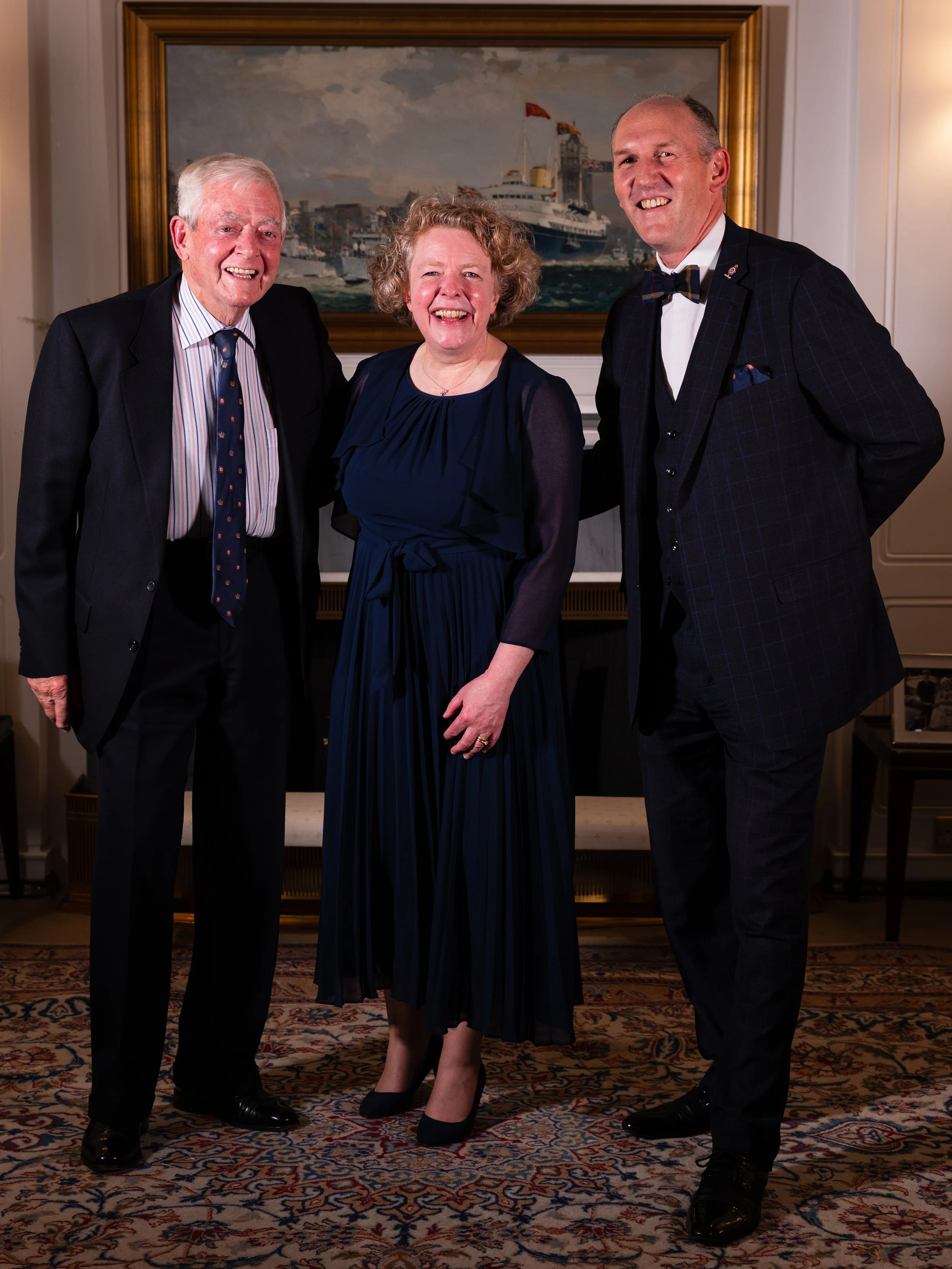 Three people standing together indoors, all dressed in formal attire, smiling at the camera with a framed maritime painting behind them.