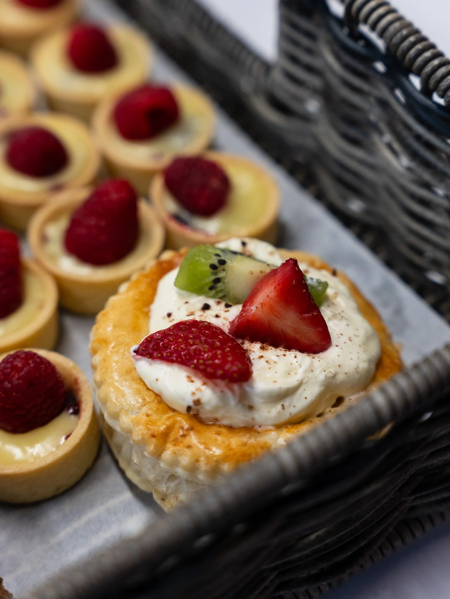 Close-up of a wicker basket filled with assorted mini cheesecakes topped with strawberries, raspberries, and kiwi slices, on a piece of parchment paper.