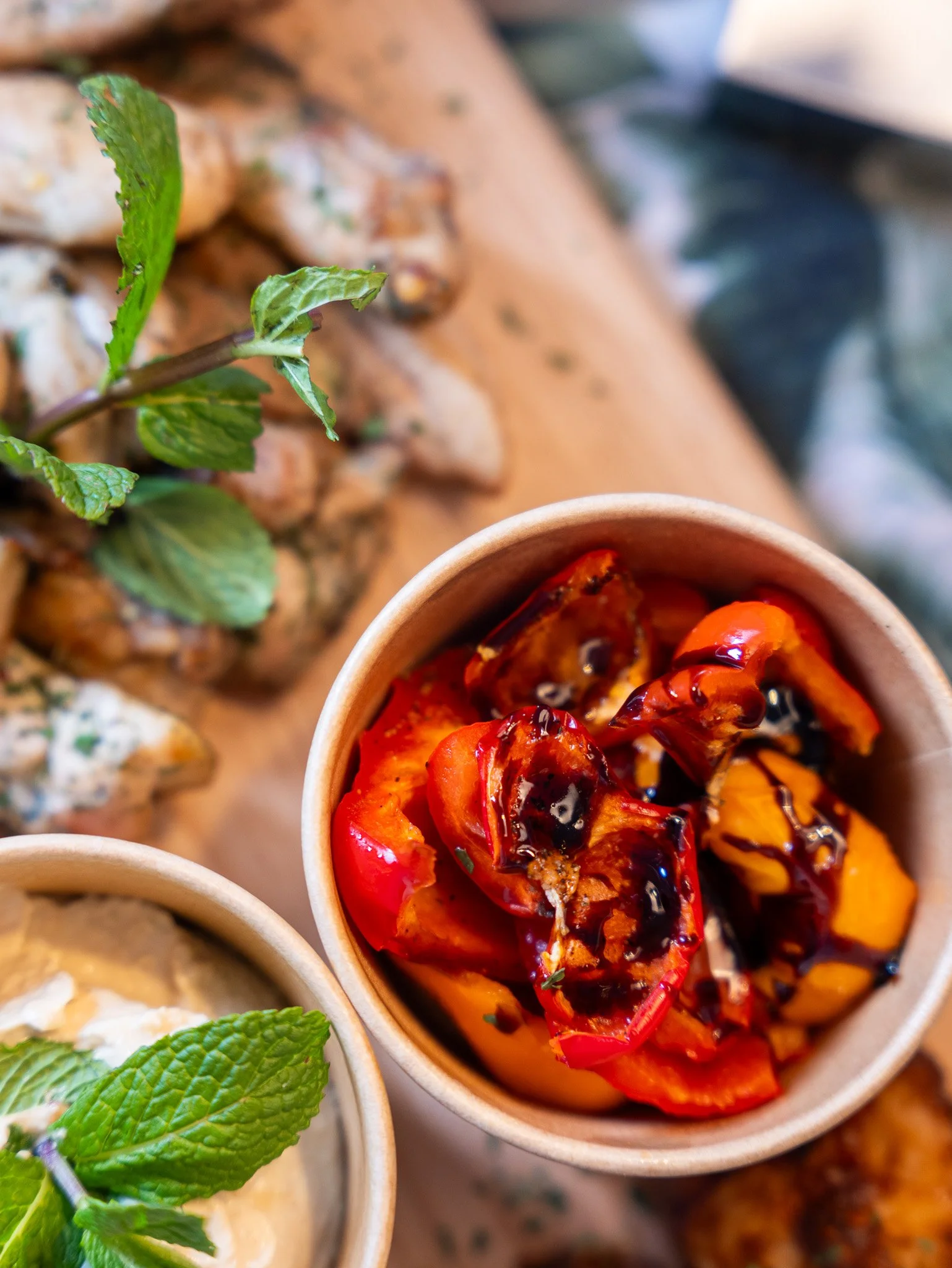 Close-up of roasted red and yellow peppers drizzled with balsamic glaze in a paper cup, with herb-marinated chicken pieces and fresh mint leaves on a wooden board in the background.