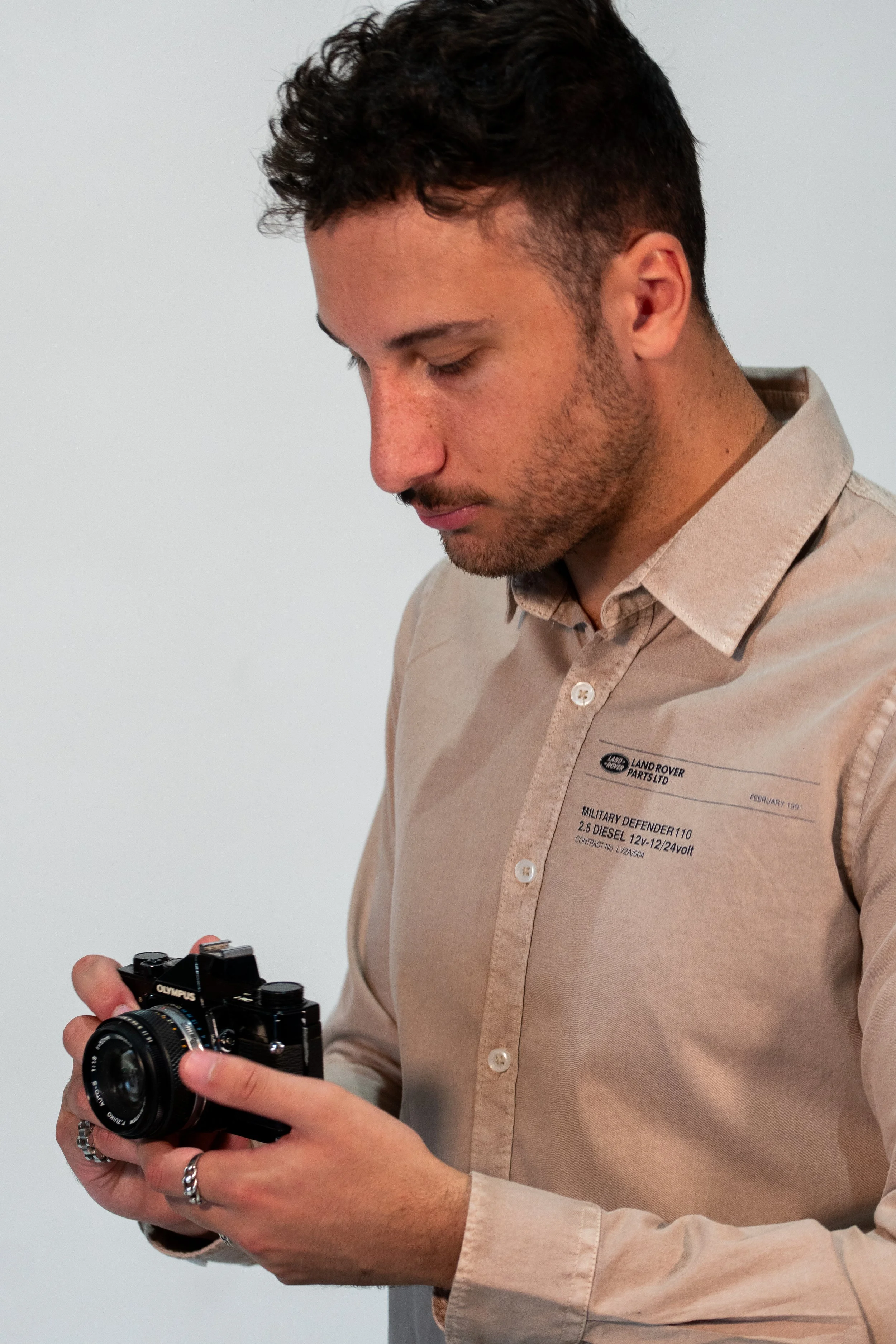 A young man with dark hair and a beard, wearing a beige shirt with text and logos, is looking down at a black Olympus camera he is holding in his hands.
