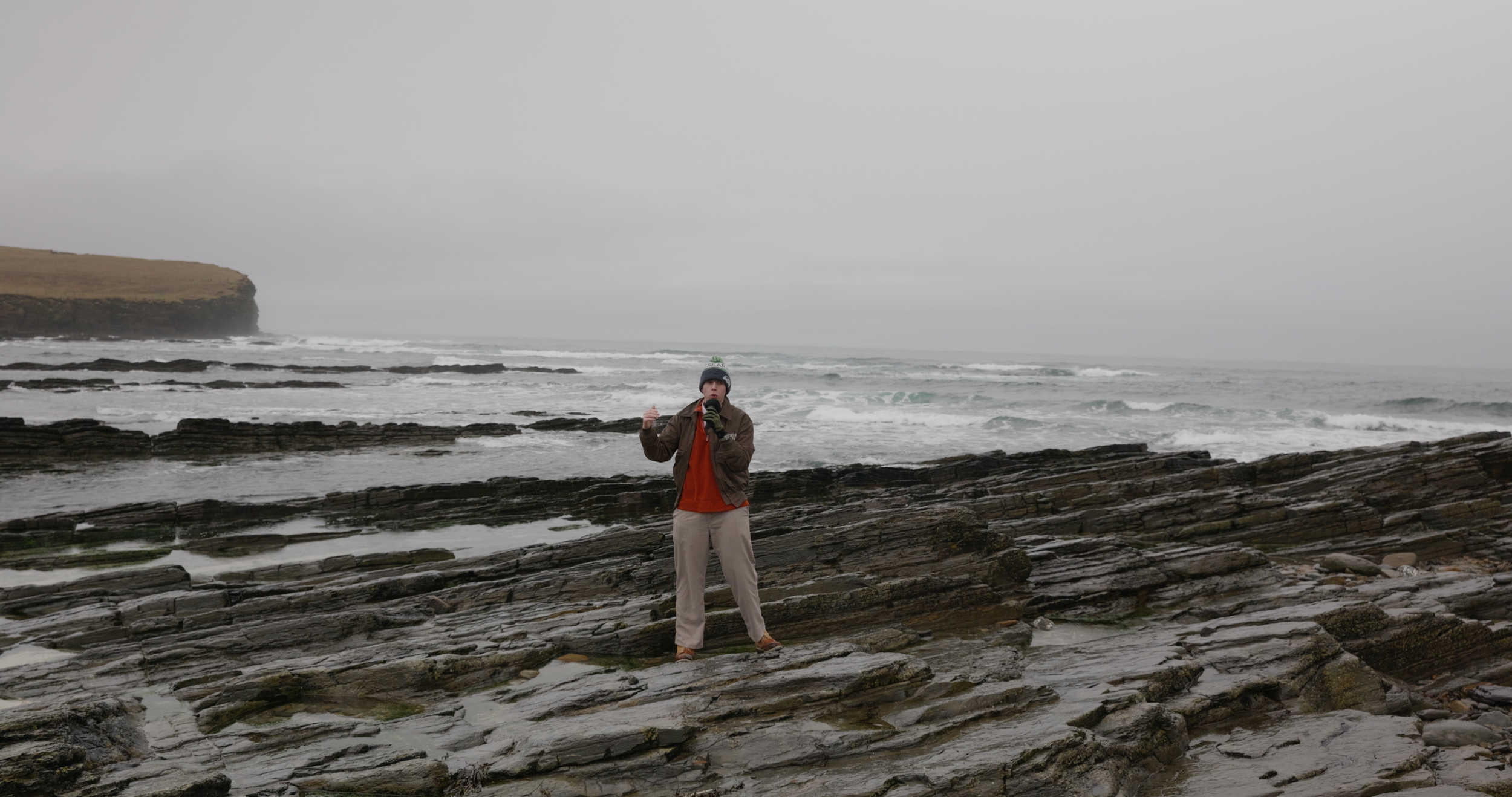 Person standing on rocky shoreline near the ocean, holding a microphone, wearing a brown jacket, orange shirt, and gray pants, with a gray beanie and gloves, under overcast skies.