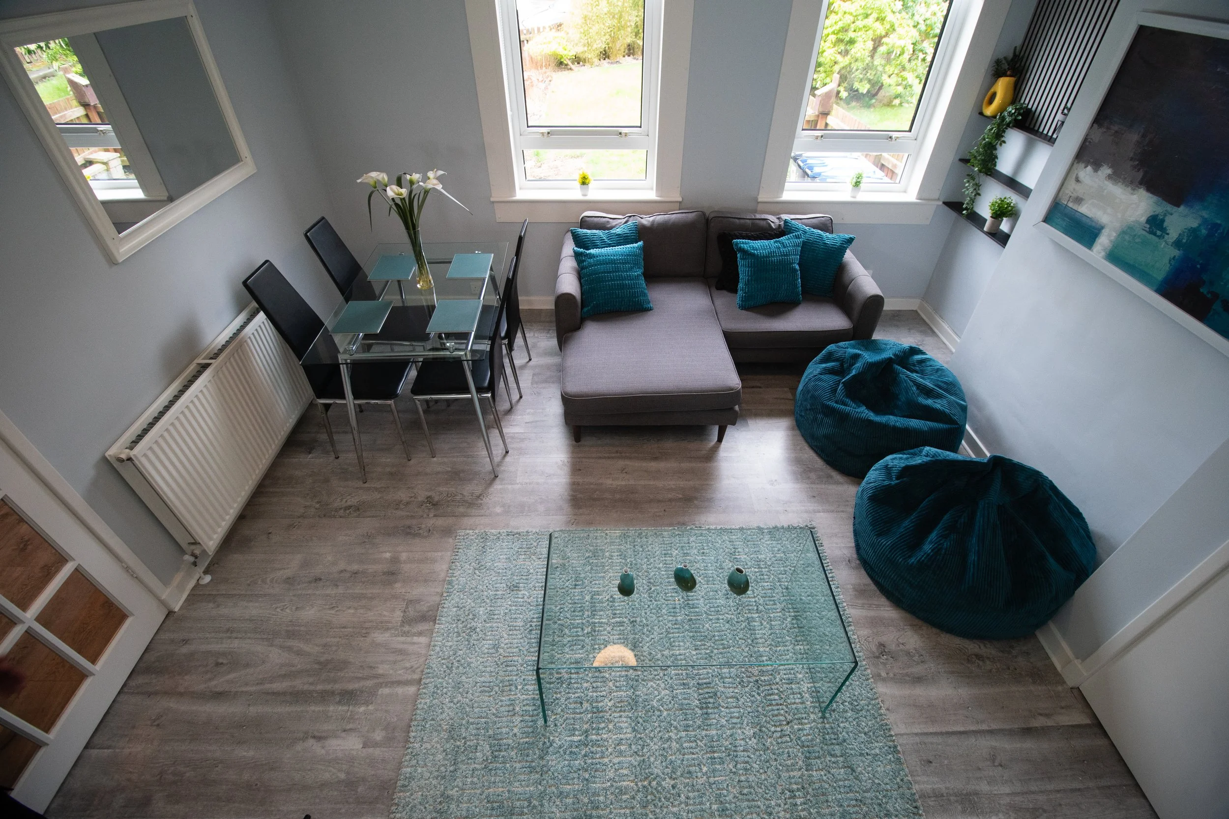 Living room with a gray sectional sofa, teal throw pillows, two teal bean bag chairs, a glass coffee table with three small vases, a dining table with four black chairs and a vase of white lilies, and large windows with a view outside.