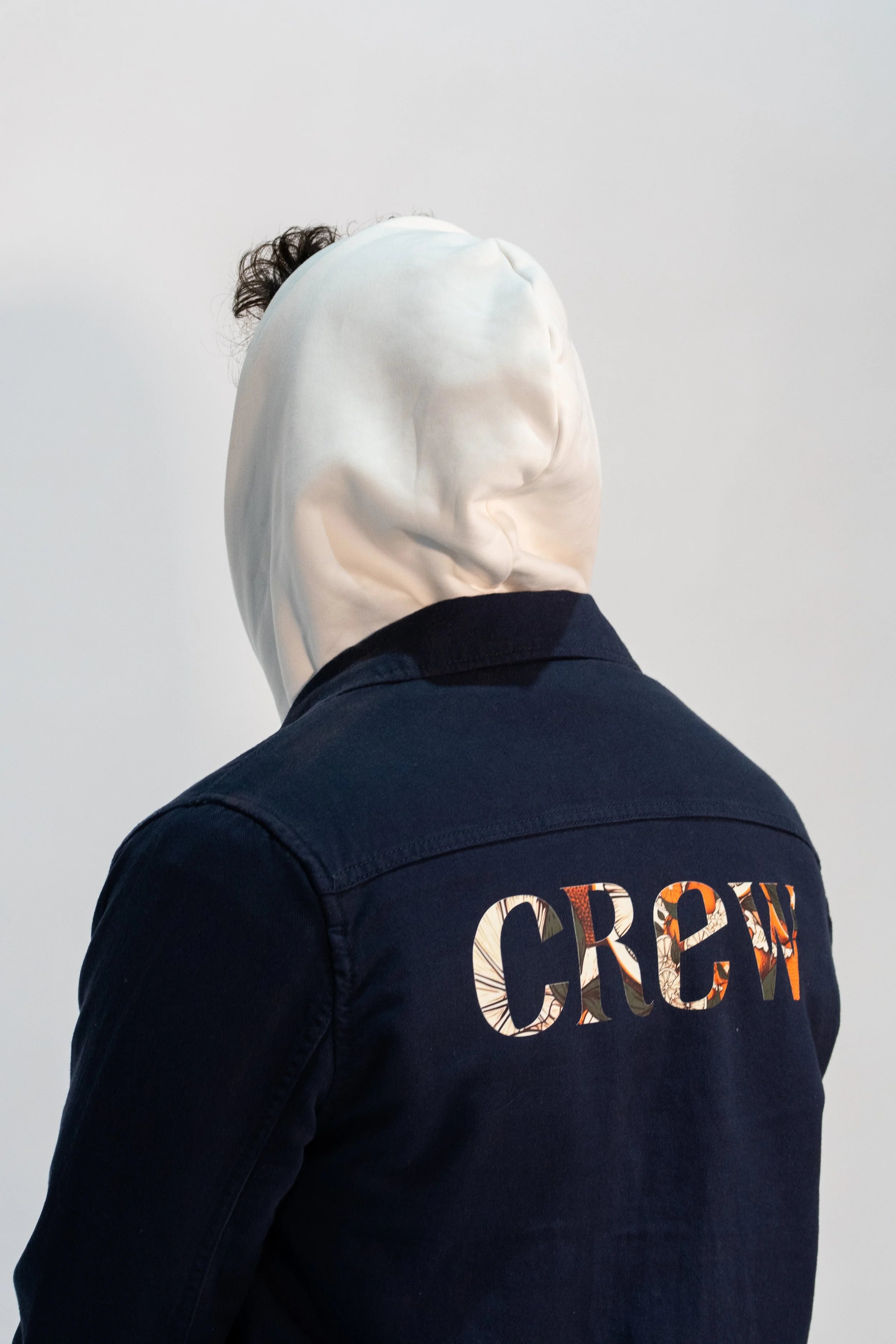 Person wearing a dark jacket with the word "CROWN" in colorful, patterned letters on the back, and a white hood covering their head, obscuring their face.