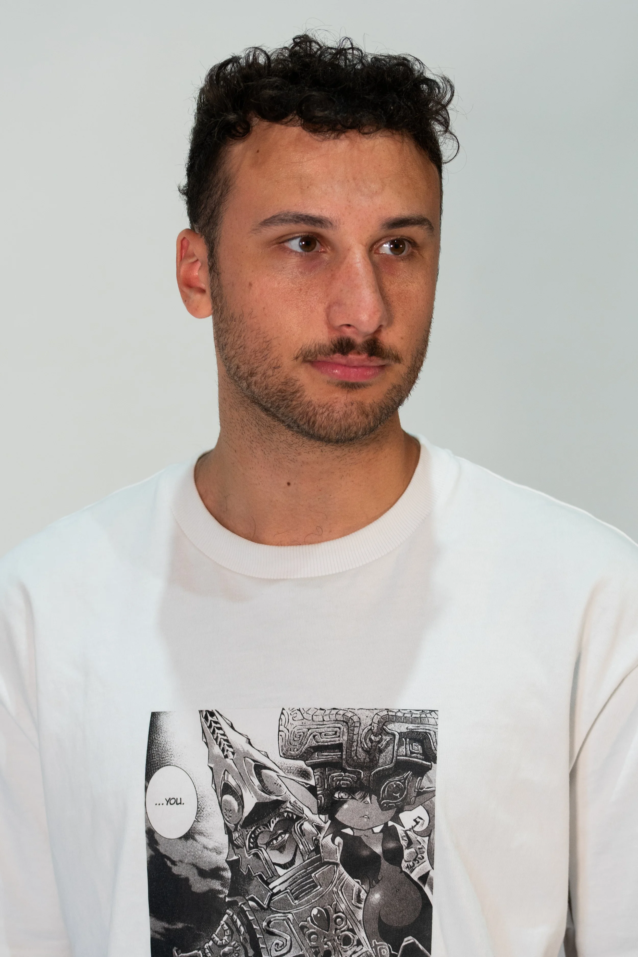 A young man with curly dark hair and a beard wearing a white T-shirt with a manga-style graphic print on the front.