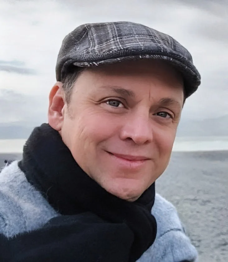 A smiling man wearing a flat cap, scarf, and coat outdoors with a cloudy sky and water in the background.