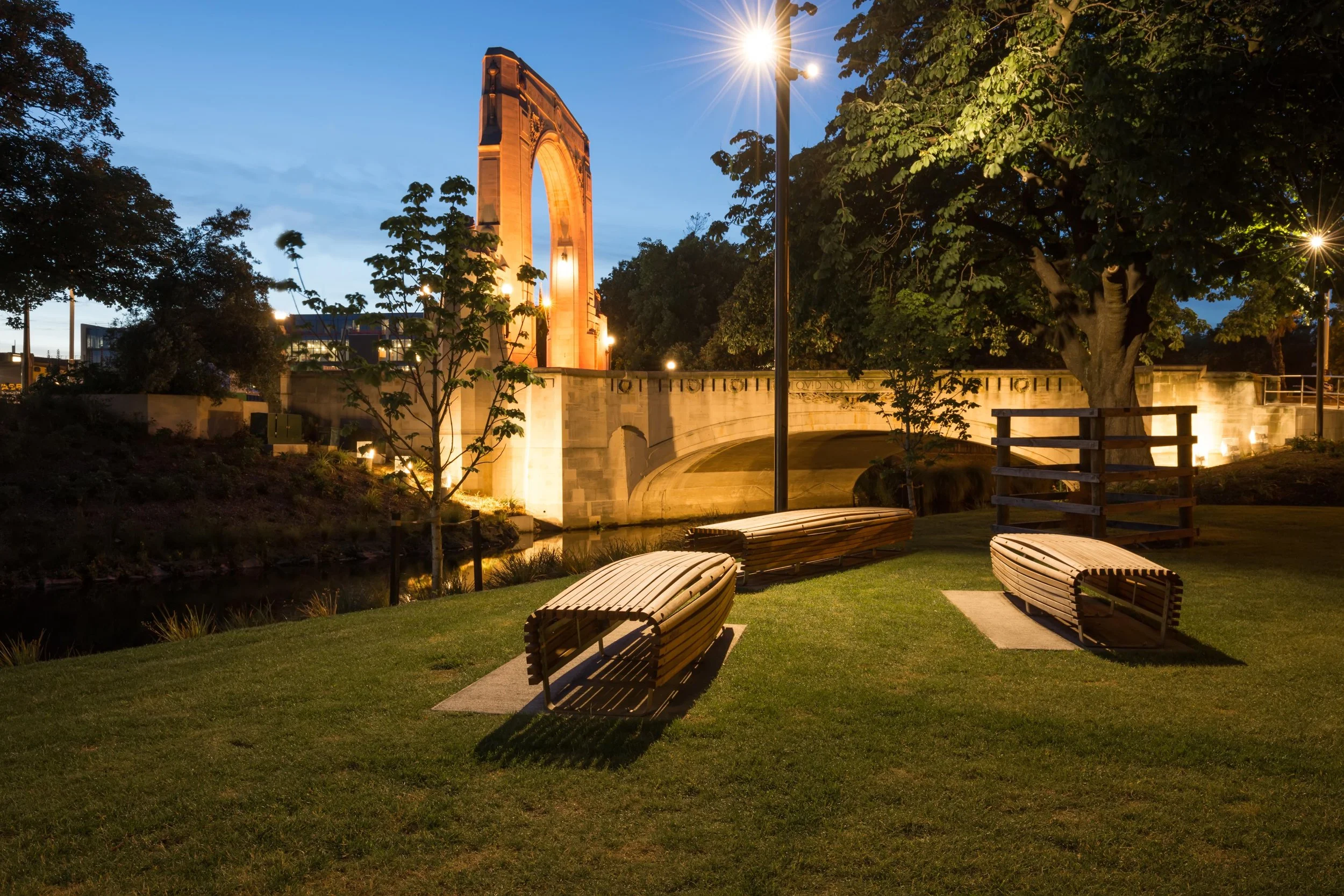 A park at dusk featuring a large arched monument illuminated with warm lights, surrounded by trees and seating benches, with streetlights and a blue sky overhead.