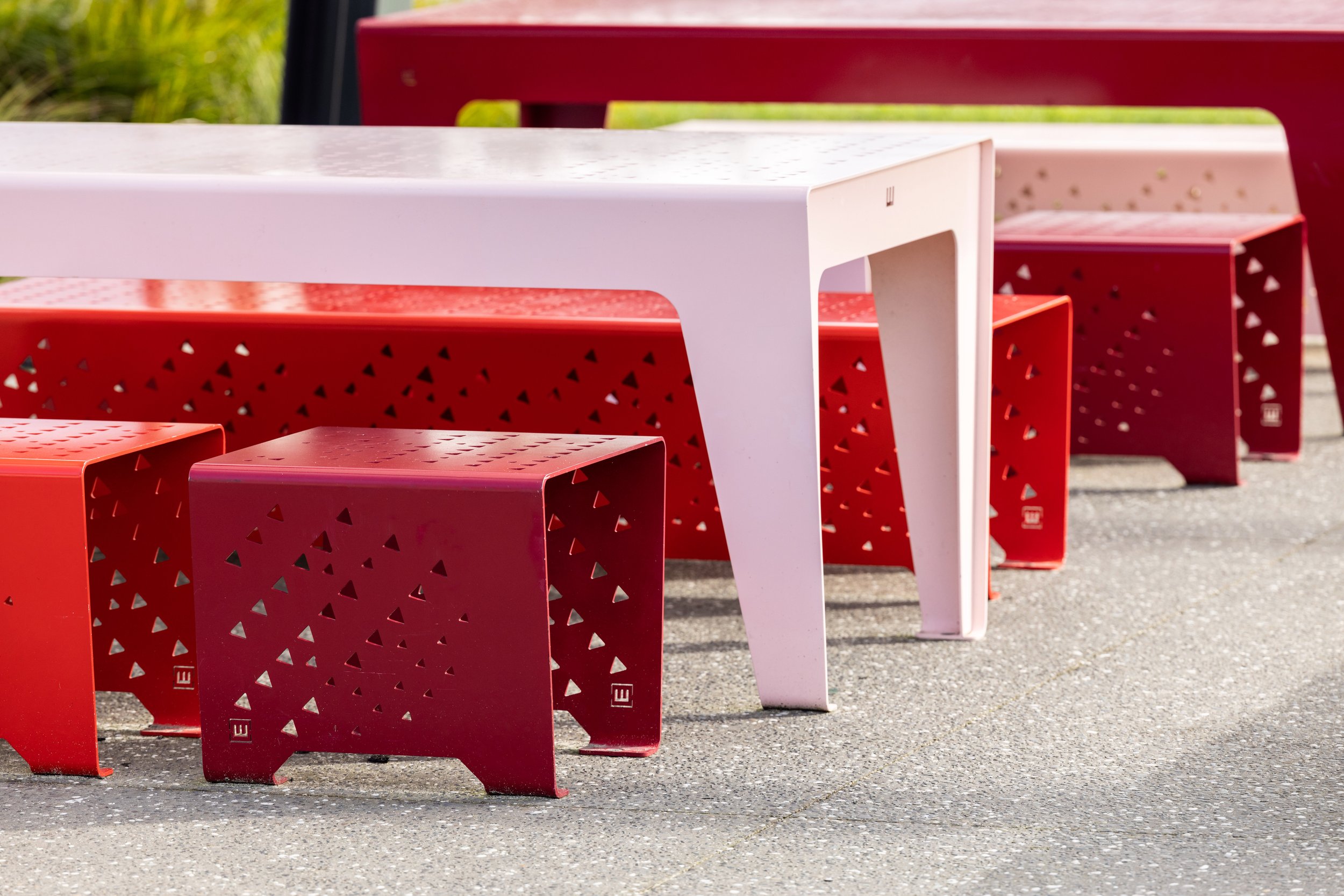 A white table and red chairs with triangular cutouts, set outdoors on a paved surface.