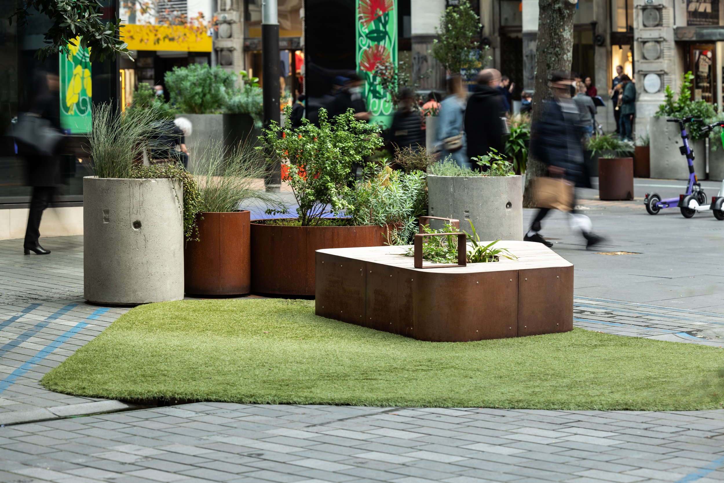 A city sidewalk with large concrete and rust-colored planters filled with green plants. Some planters are placed on a square patch of artificial grass. There are blurred pedestrians walking past, and storefronts with colorful signs and displays in the background.