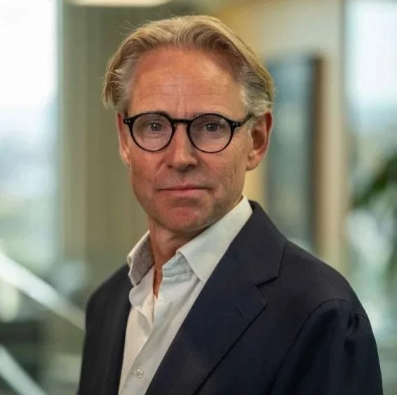 En man i formell kostym och glasögon står i ett kontorsomrade.