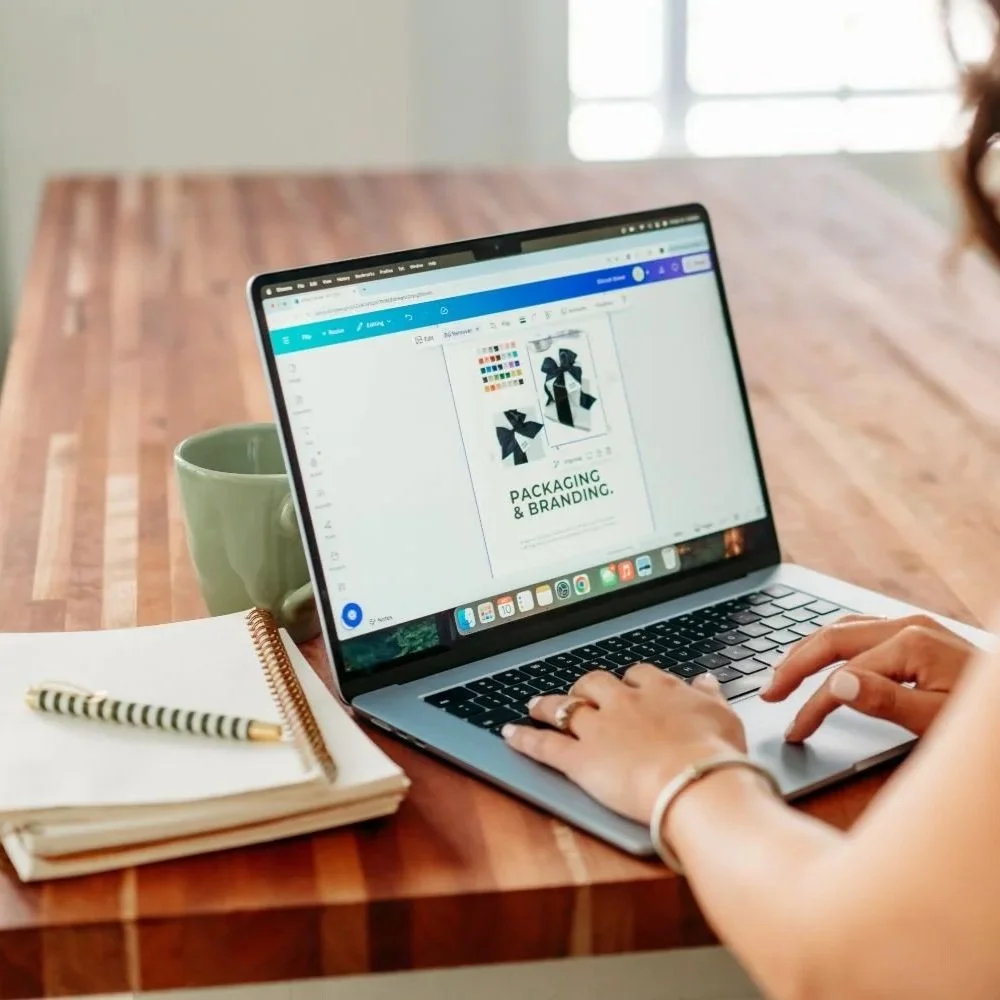 A woman working on a laptop at a wooden table with a notebook and a green mug nearby.