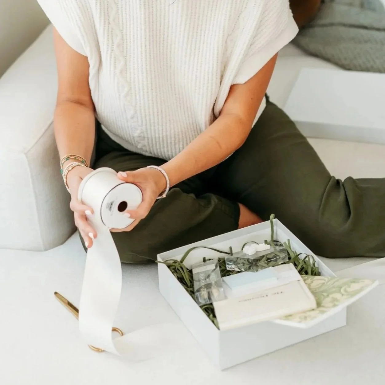 A woman sits on a couch, holding a ribbon spool,  working on assembling a gift box.