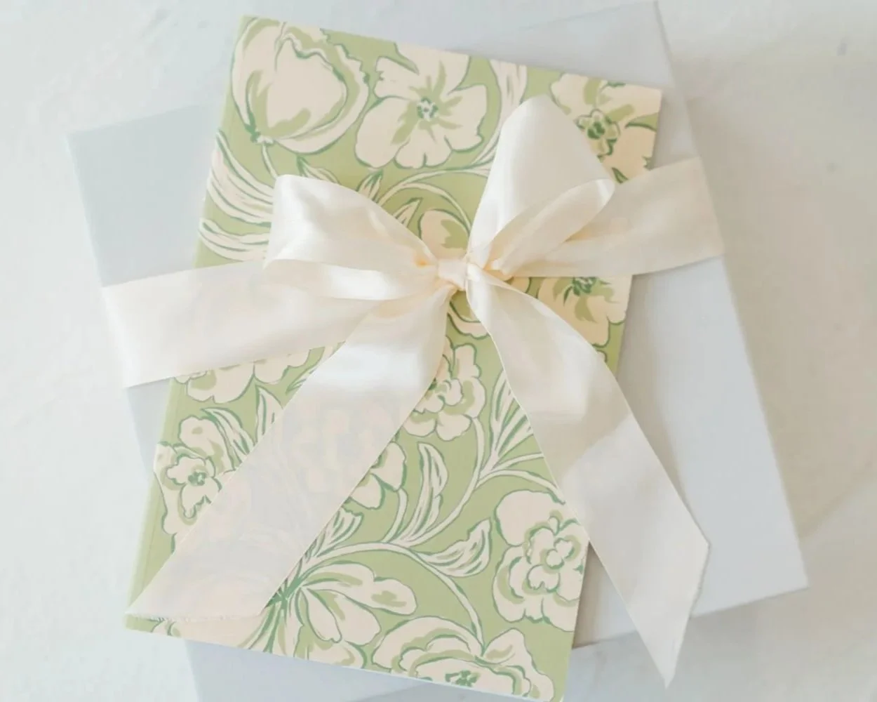 A gift box wrapped in floral-patterned green and white paper, adorned with a large white ribbon tied into a bow.