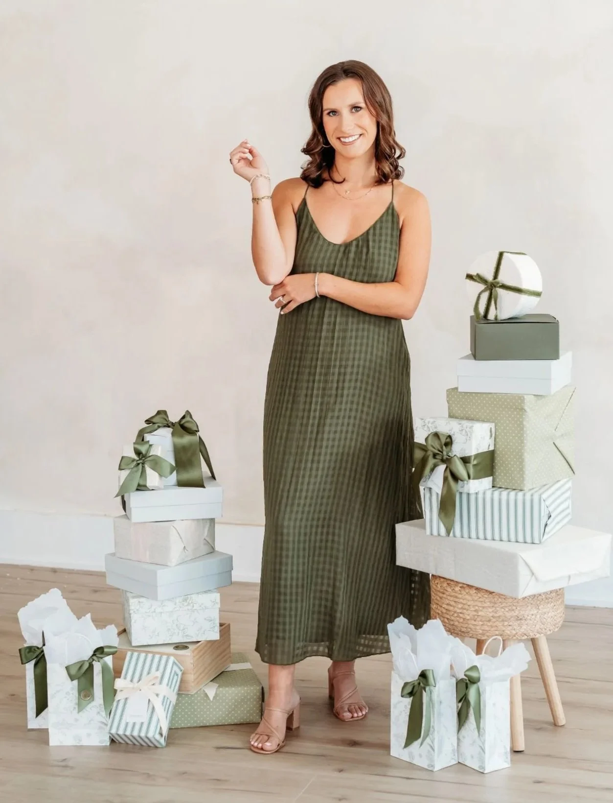A woman in a long dark dress smiling and standing among neatly wrapped gifts with green ribbons against a soft gray background.