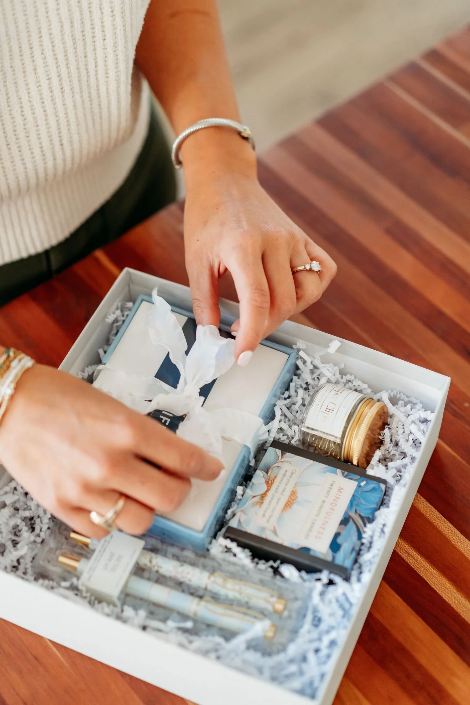 A woman unwrapping a gift box filled with shredded paper, cosmetics, and small jars. The woman's hands are adorned with rings.