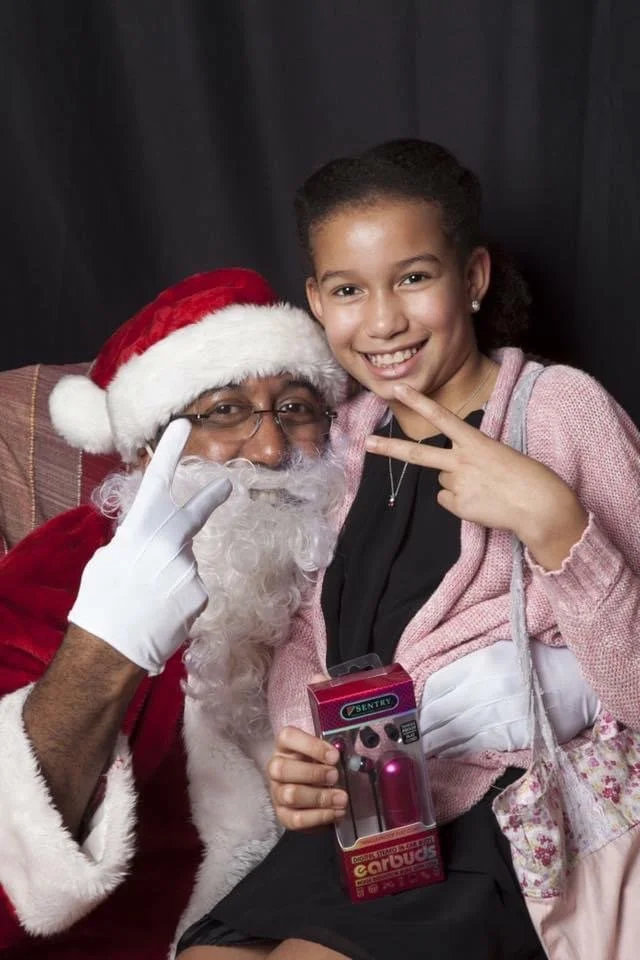 A young girl sitting with Santa Claus, who is wearing a red suit, a Santa hat, glasses, and a white beard. The girl is smiling and making a peace sign with her right hand, holding a small pink and purple toy in her left hand. She is wearing a pink sweater over a black top. The background is dark.